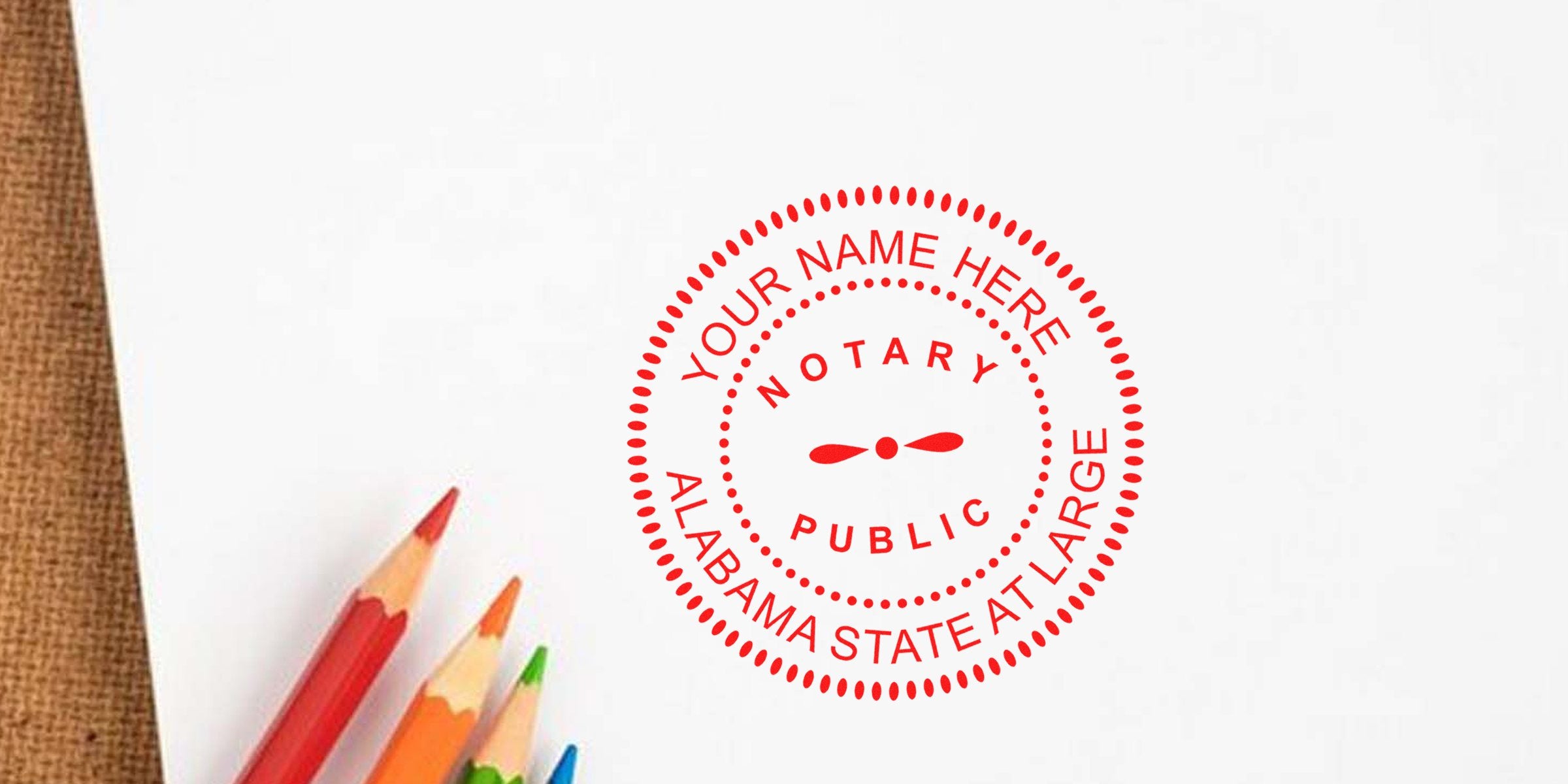 Red circular notary public stamp reading "Your Name Here, Alabama State At Large" on white paper with colorful pencils and burlap edge