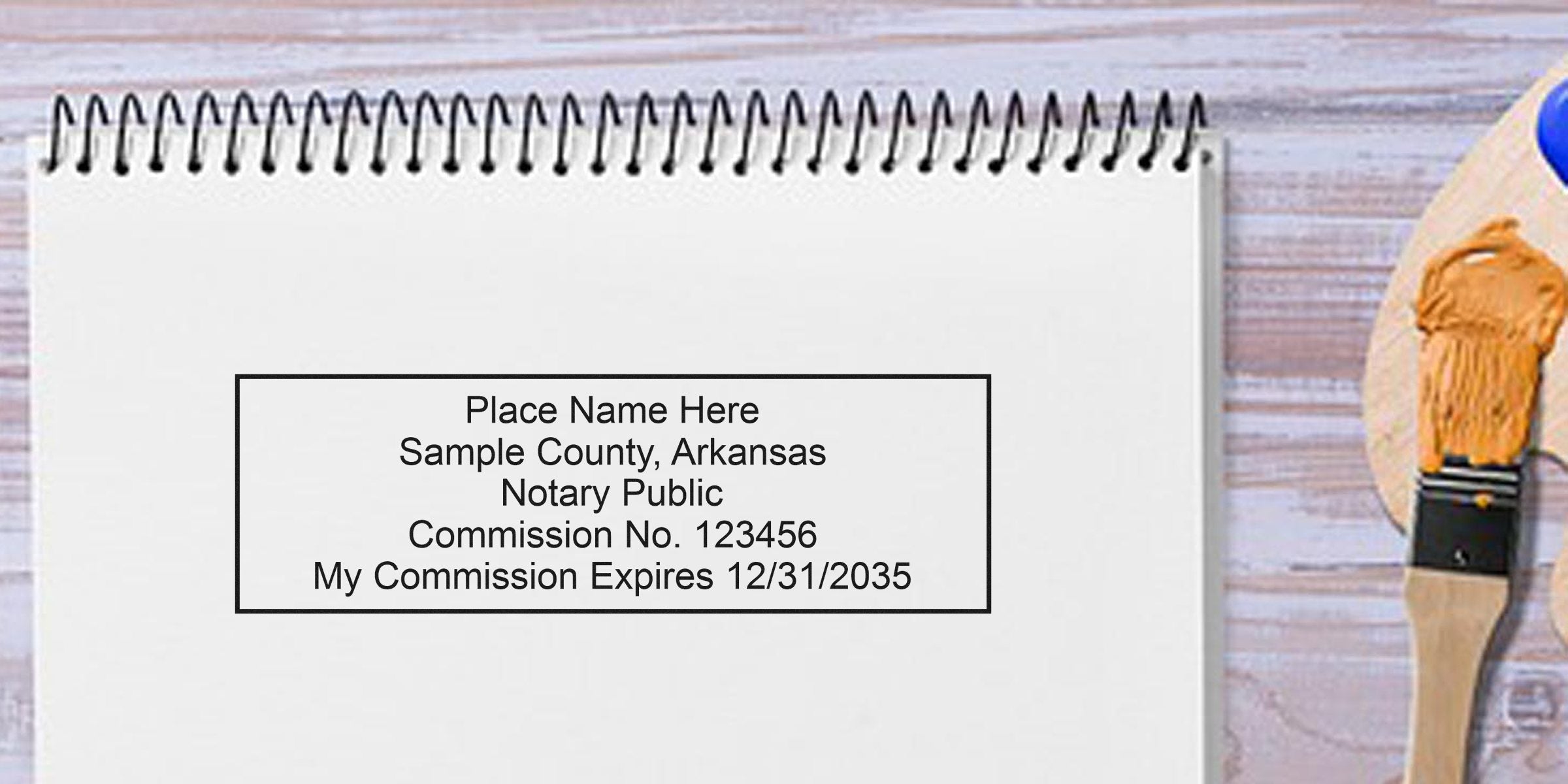 Mock notary public stamp sample on spiral notebook reading "Place Name Here, Sample County, Arkansas, Notary Public, Commission No. 123456, My Commission Expires 12/31/2035" with a paintbrush on a wooden table background