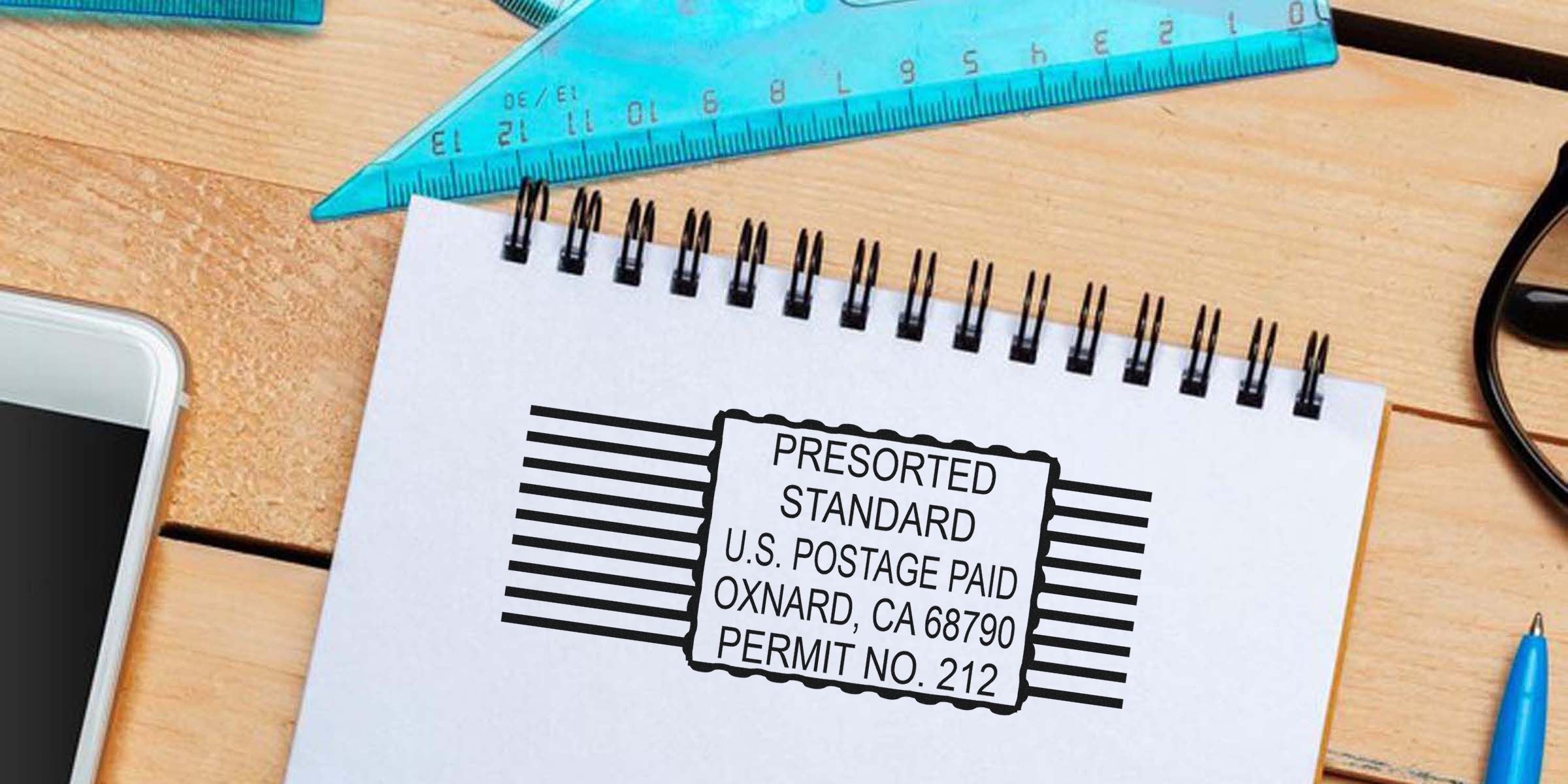 A notebook page with a barcode stamp reading “PRESORTED STANDARD U.S. POSTAGE PAID OXNARD, CA 68790 PERMIT NO. 212” on a wooden desk, beside a ruler, phone, glasses and a blue pen.