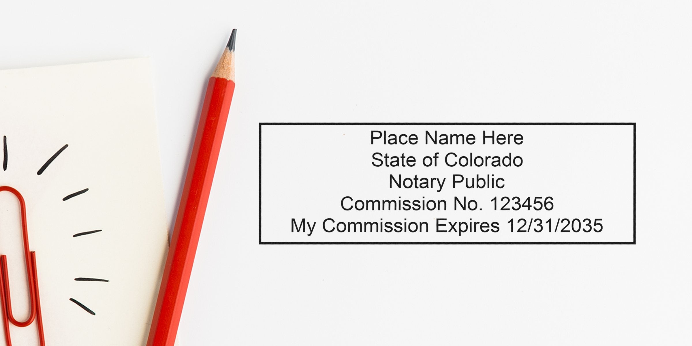 Top-down photo of a notary stamp template reading "Place Name Here, State of Colorado, Notary Public, Commission No. 123456, My Commission Expires 12/31/2035" beside a red pencil and red paperclip on a white background