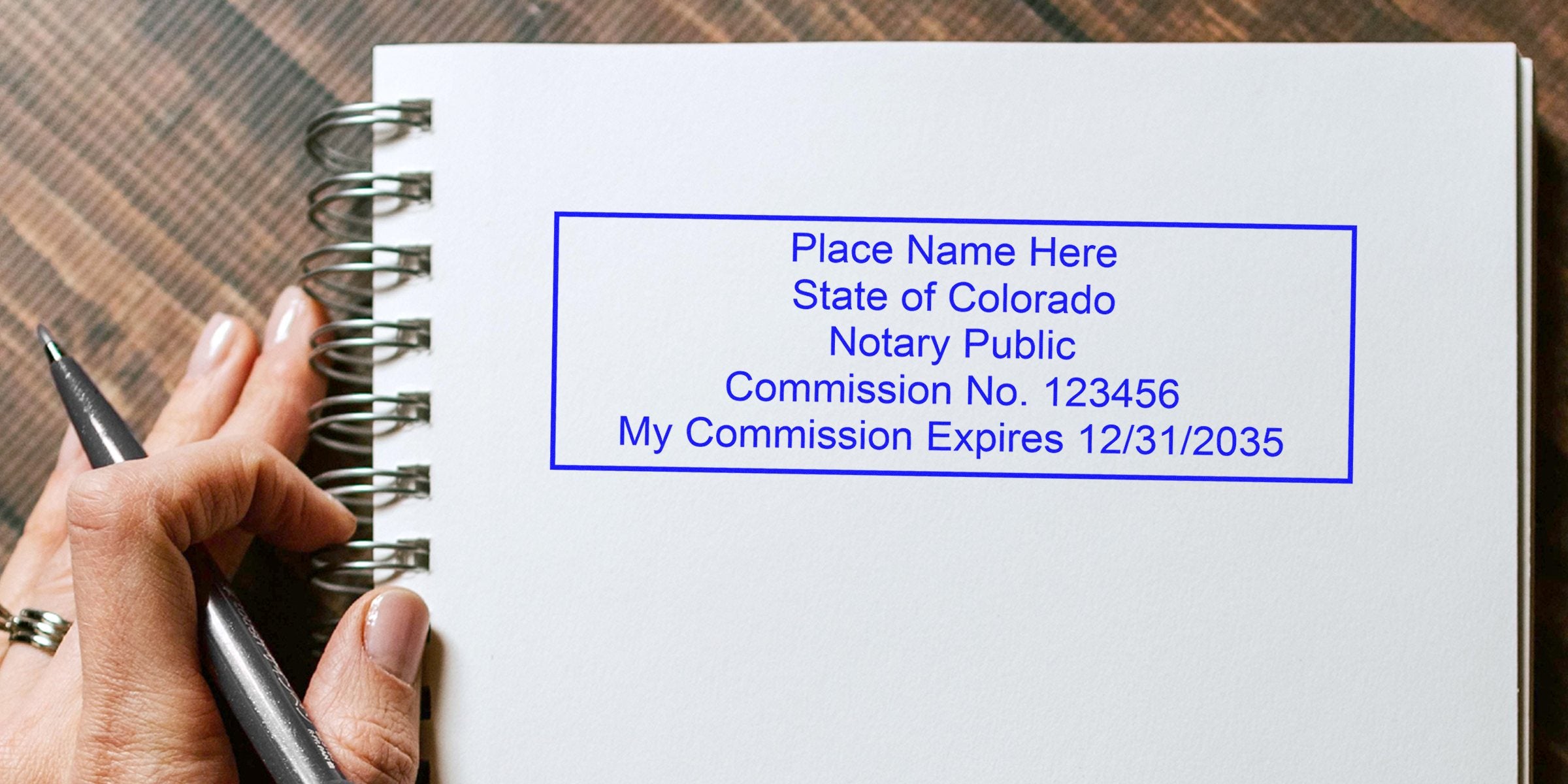 Blue notary stamp mockup on white spiral notebook reading "State of Colorado Notary Public, Commission No. 123456, Expires 12/31/2035" with a hand holding a pen.