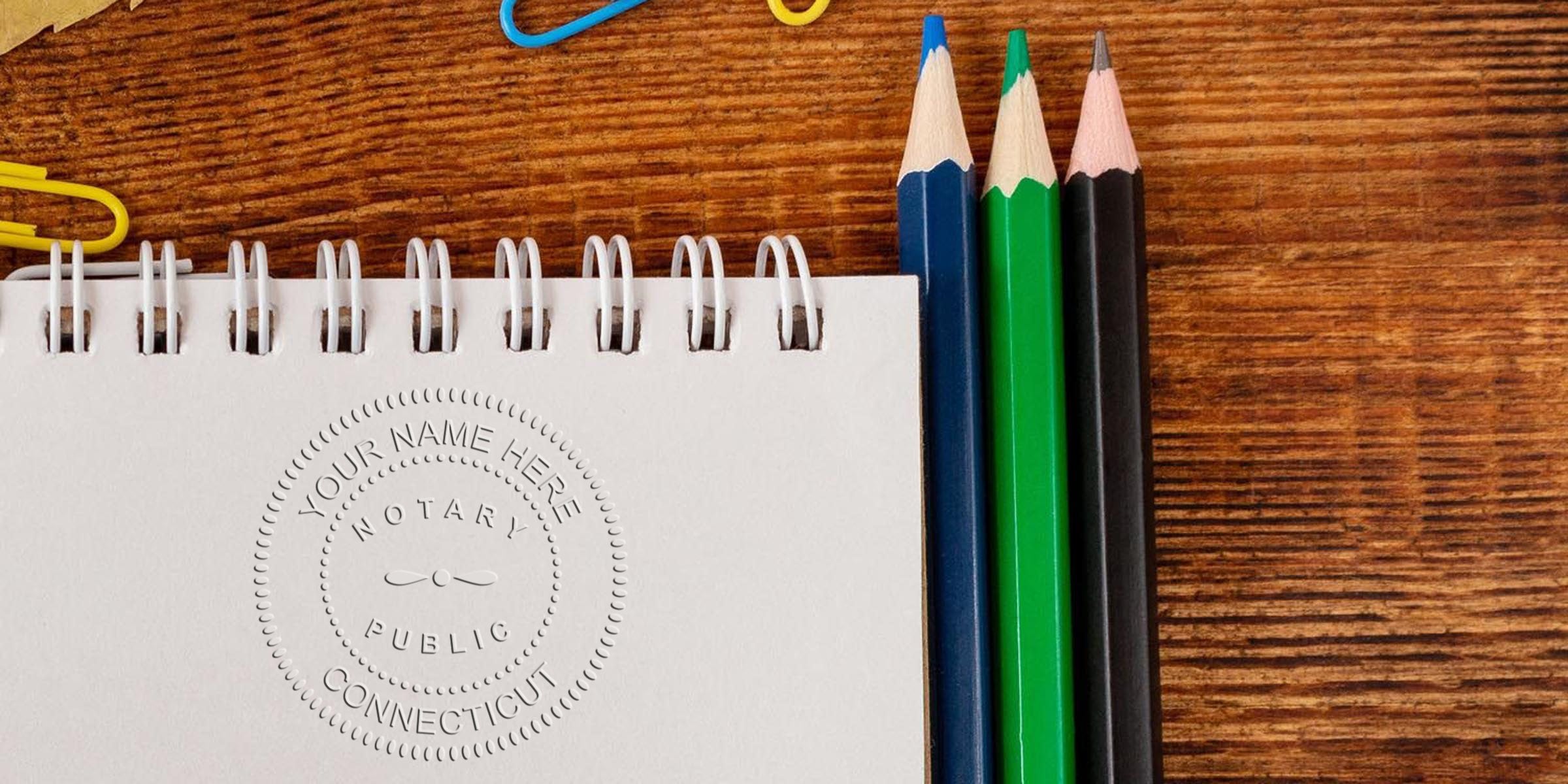 Close-up of embossed Notary Public Connecticut seal on spiral notebook page with blue, green and black colored pencils and paperclips on a textured wooden desk surface