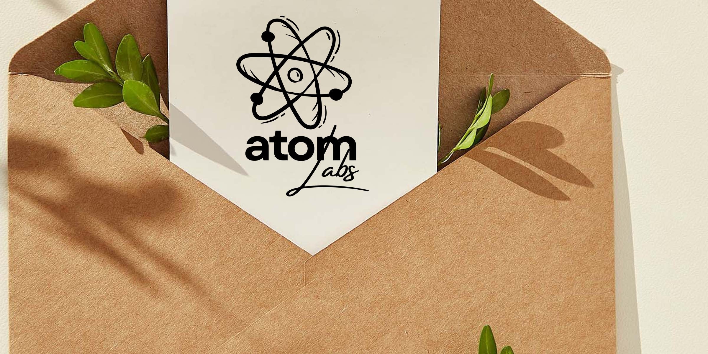 Kraft paper envelope with a white card showing a black atom logo and "Atom Labs" text, green foliage tucked inside and soft shadows on a neutral background.