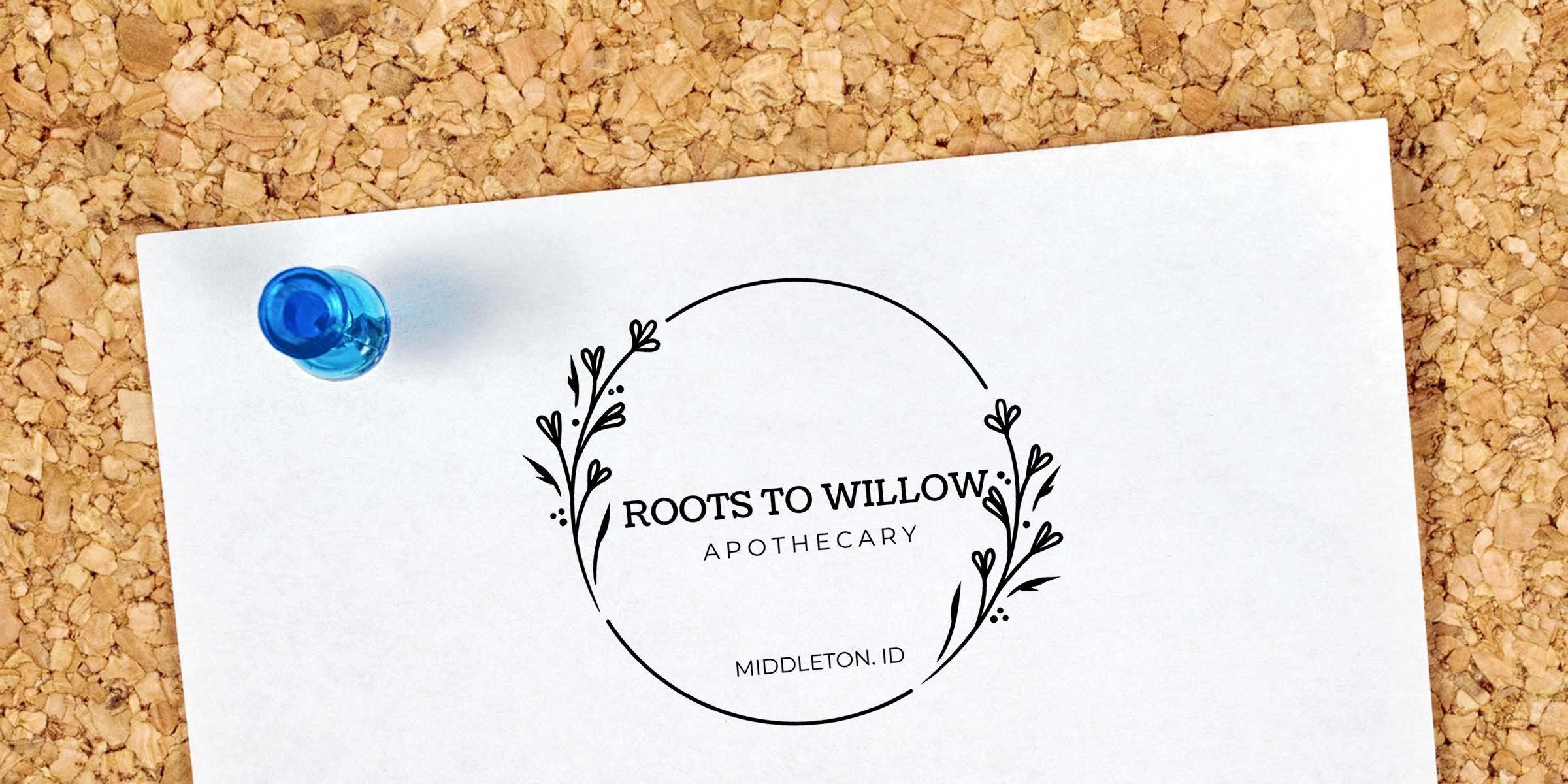 White paper pinned to corkboard with a blue pushpin displaying a minimalist botanical wreath logo reading "Roots to Willow Apothecary, Middleton ID" — apothecary branding, herbal shop logo.