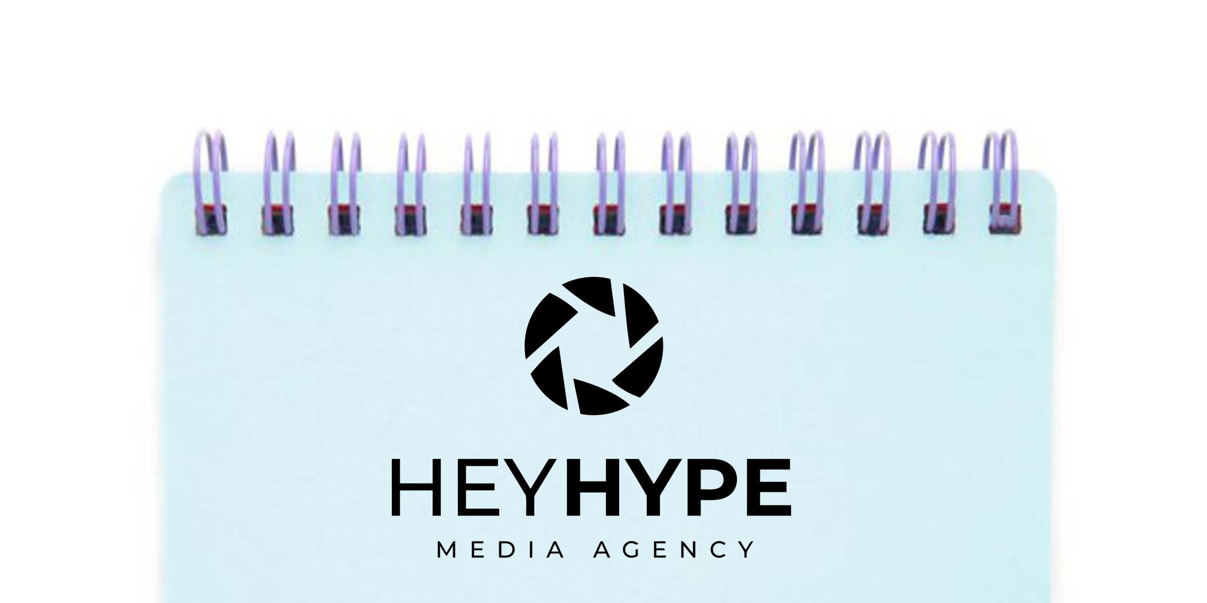 Light blue spiral-bound notebook top showing a black camera aperture logo above bold "HEYHYPE" and smaller "MEDIA AGENCY" text — minimalist media agency branding on stationery mockup.