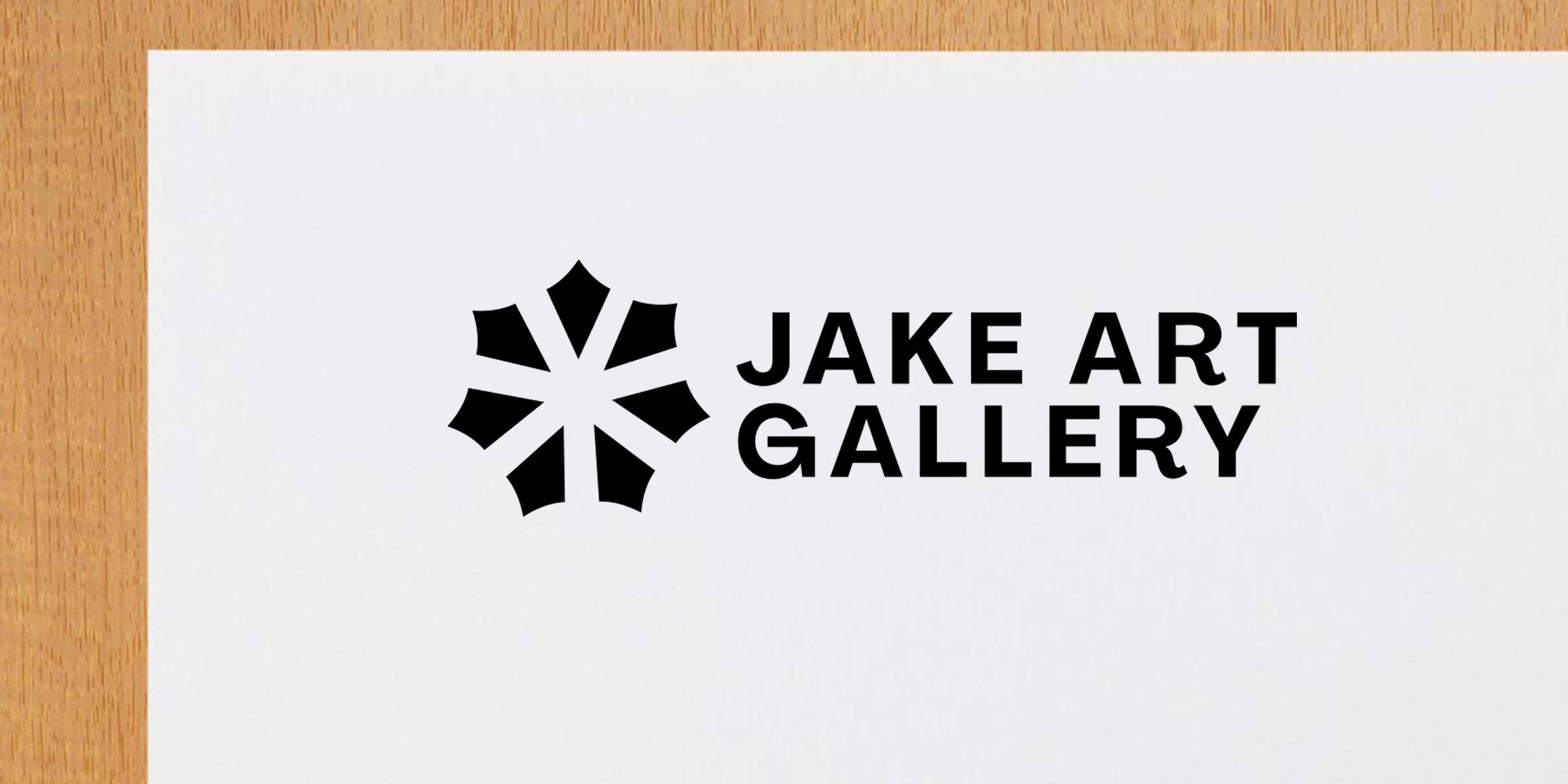 Jake Art Gallery black geometric star logo with bold uppercase wordmark on white paper against a wooden table edge, minimalist modern gallery branding