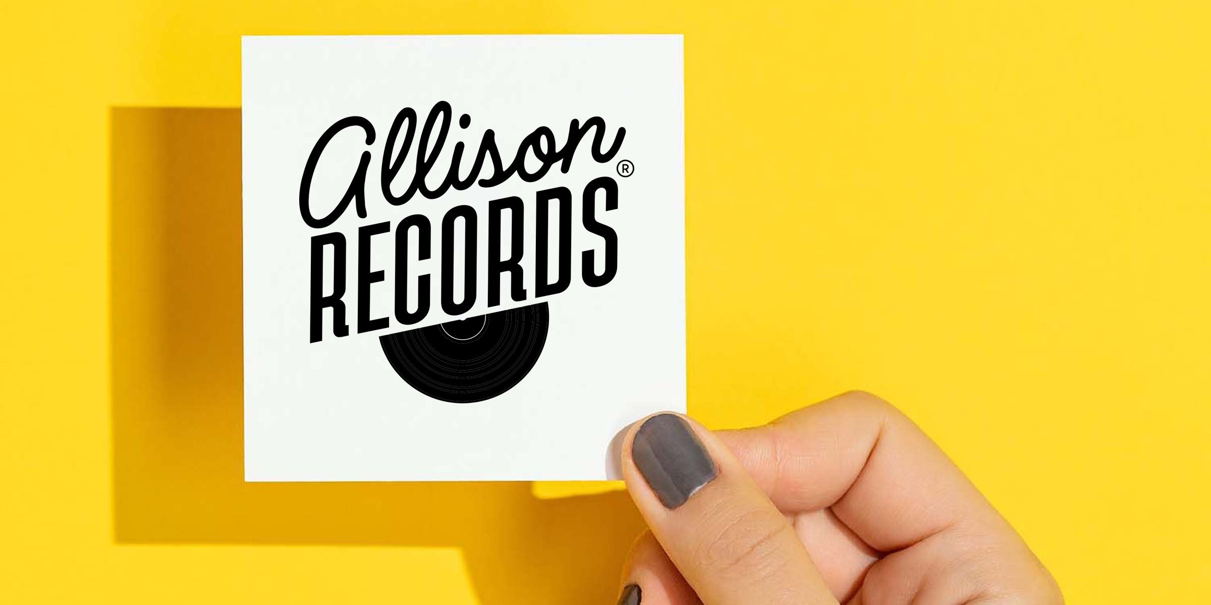 Hand holding white square card with black "Allison Records" logo and vinyl graphic against bright yellow background, close-up showing gray nail polish and card shadow for music branding and business card concept