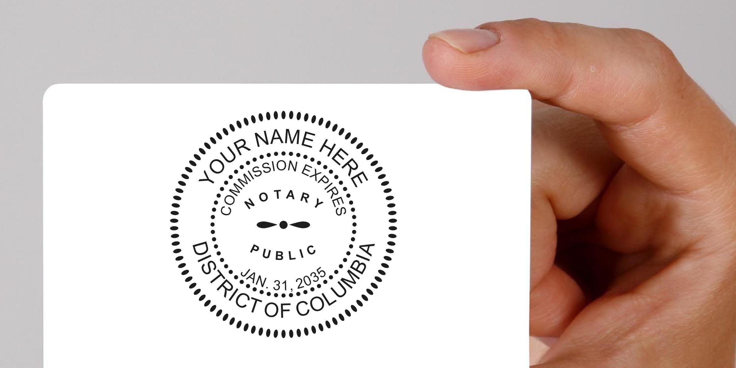 Hand holding white card with black circular notary public seal reading "Your Name Here", "Notary Public", "Commission Expires Jan. 31, 2035", "District of Columbia" — blank card showcasing a notary stamp design.