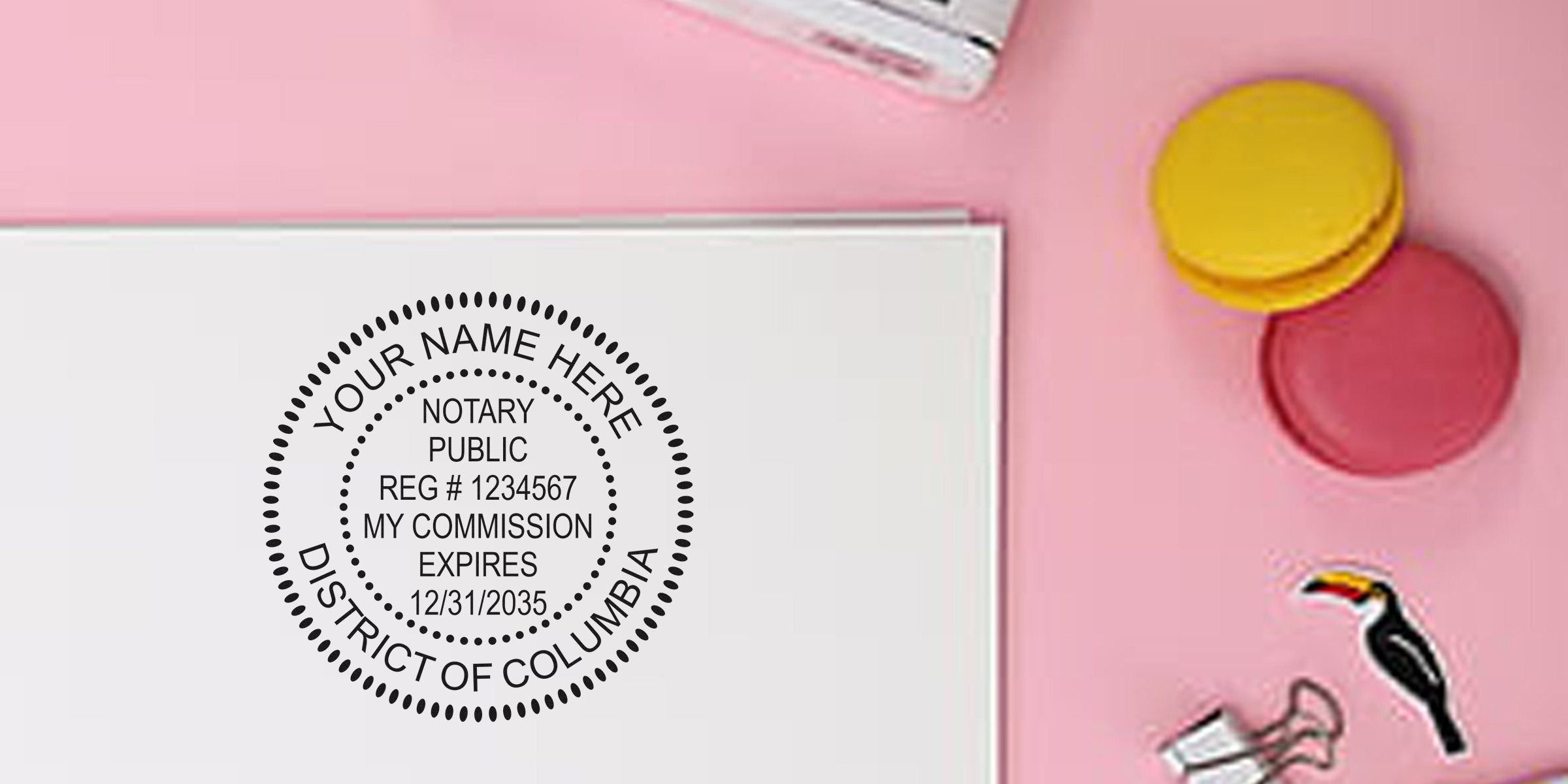 Flat lay of a personalized Notary Public seal mockup (District of Columbia) stamped on white paper on a pink desk, with colorful macarons, binder clip and toucan sticker