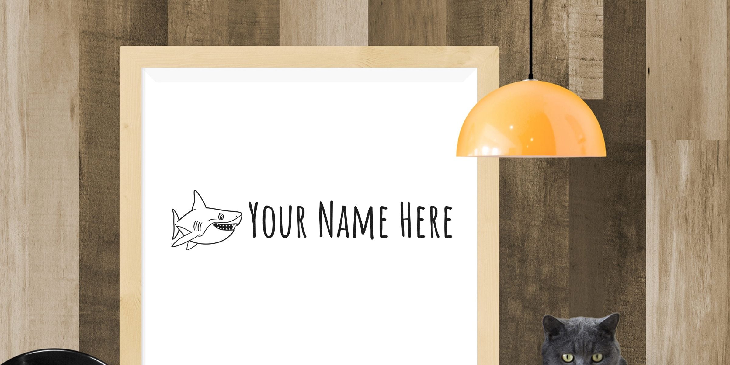 Cartoon shark graphic and the words "Your Name Here" on a whiteboard mounted on a wooden-walled background, with an orange pendant lamp overhead.