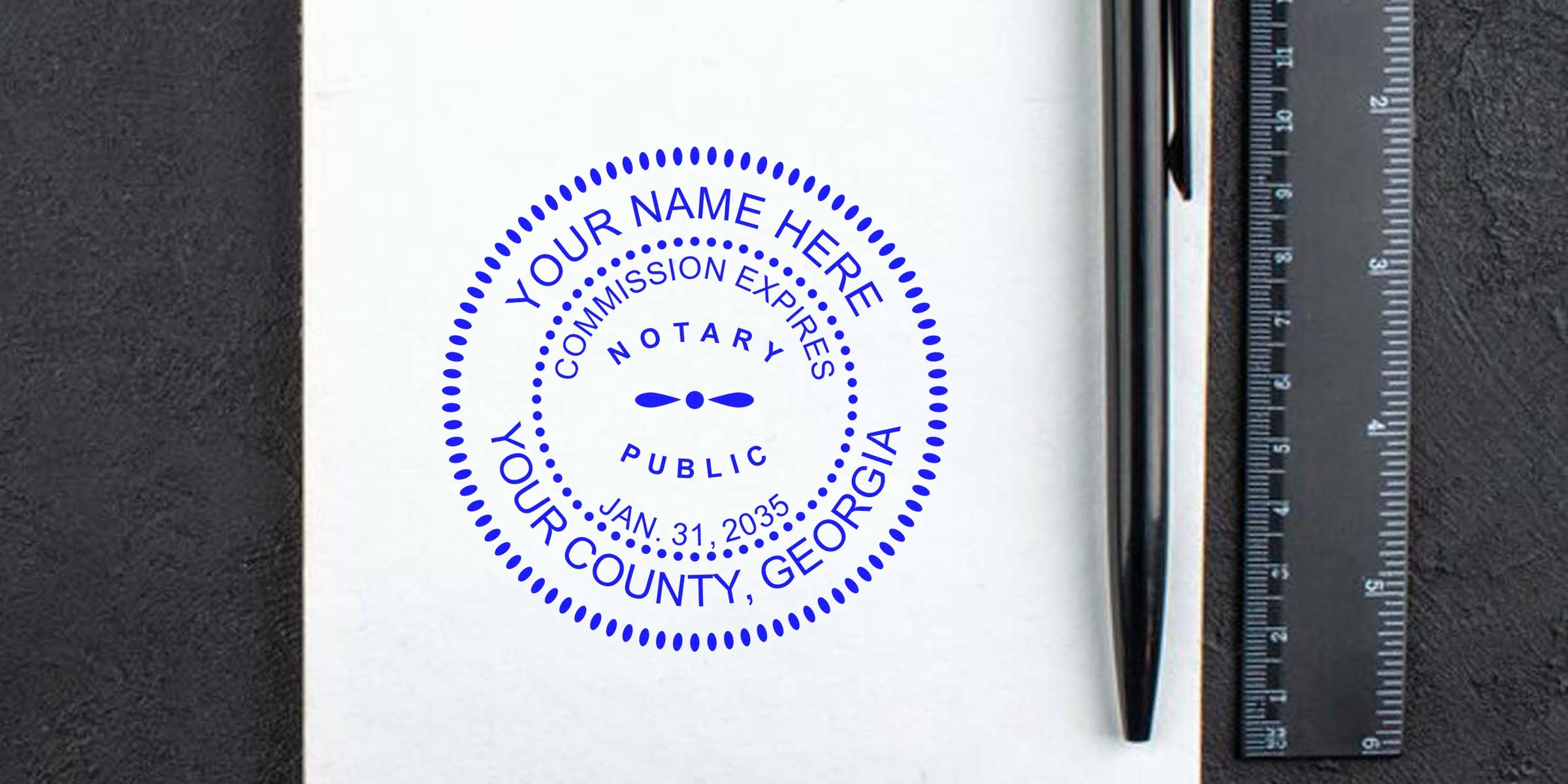 Blue circular notary public seal mockup stamped on white paper reading "Your Name Here, Your County, Georgia" with sample expiration date, placed next to a black pen and ruler on a dark textured desk — notary stamp, notary public seal, Georgia.