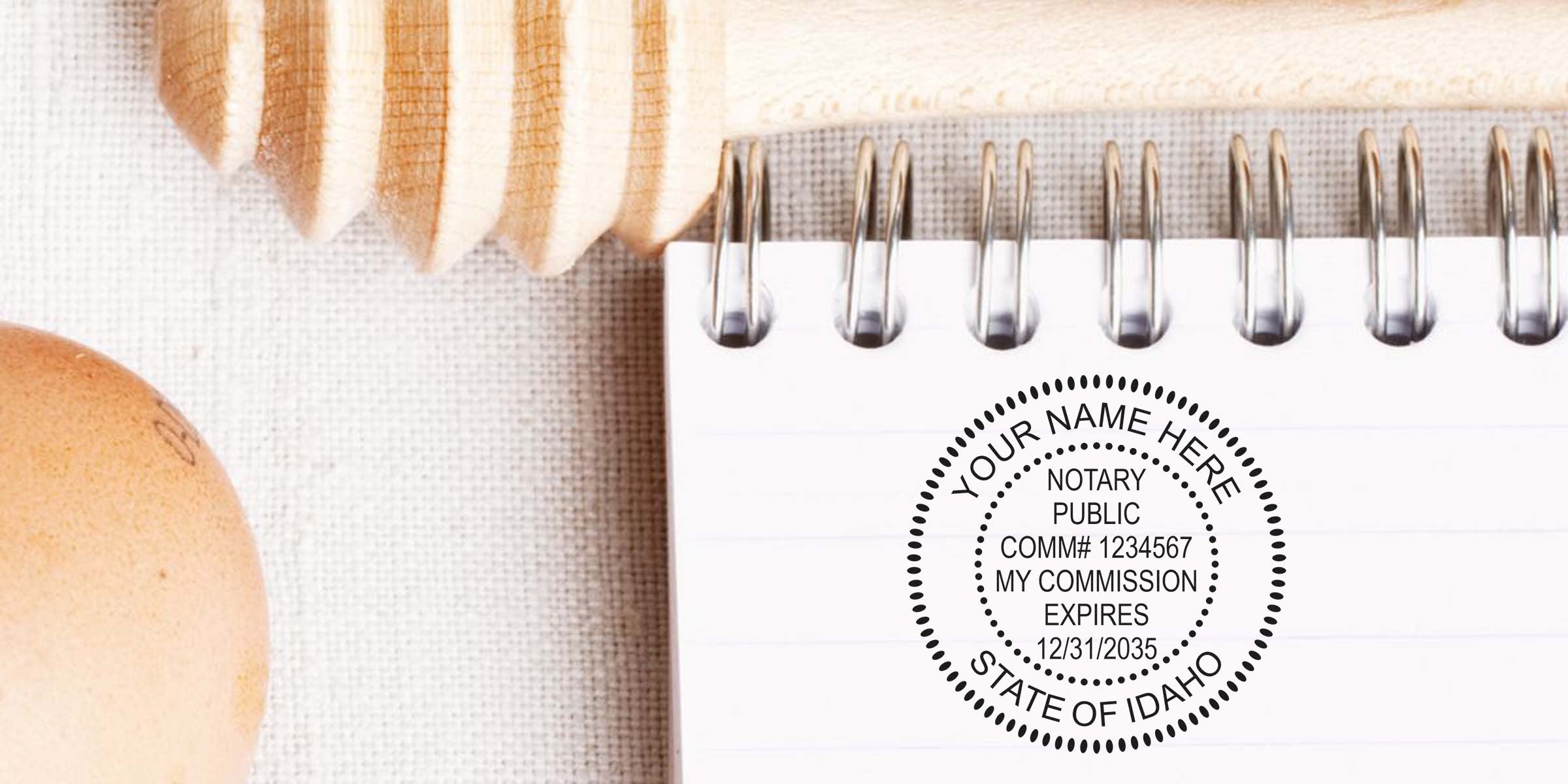 Spiral notepad with circular Idaho notary public stamp mockup reading "Your Name Here," placed beside a wooden honey dipper and brown egg on a textured fabric background.