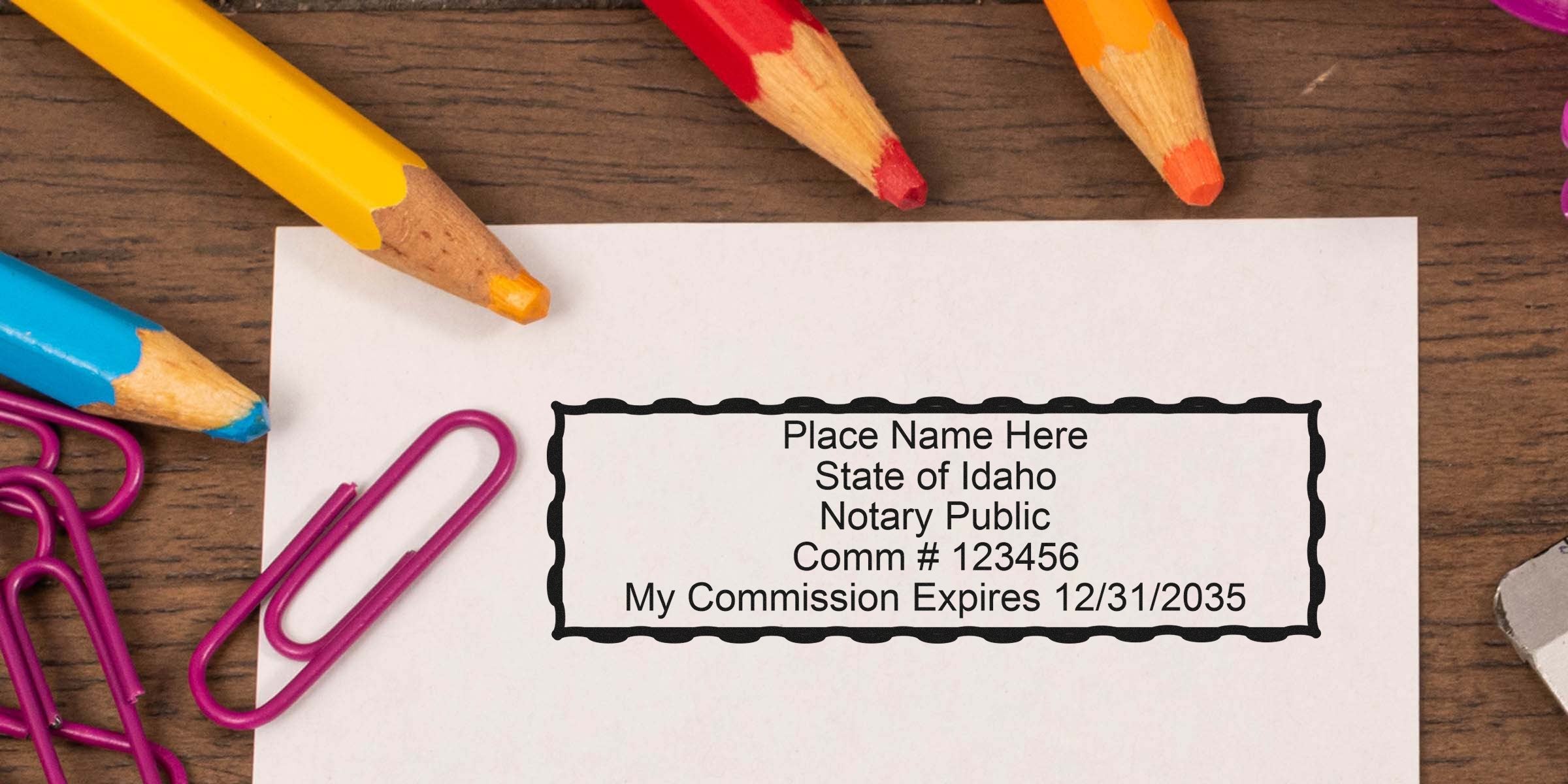 Paper with rectangular notary stamp sample reading "Place Name Here, State of Idaho, Notary Public, Comm #123456, My Commission Expires 12/31/2035" surrounded by colorful colored pencils and purple paperclips on a wooden desk.