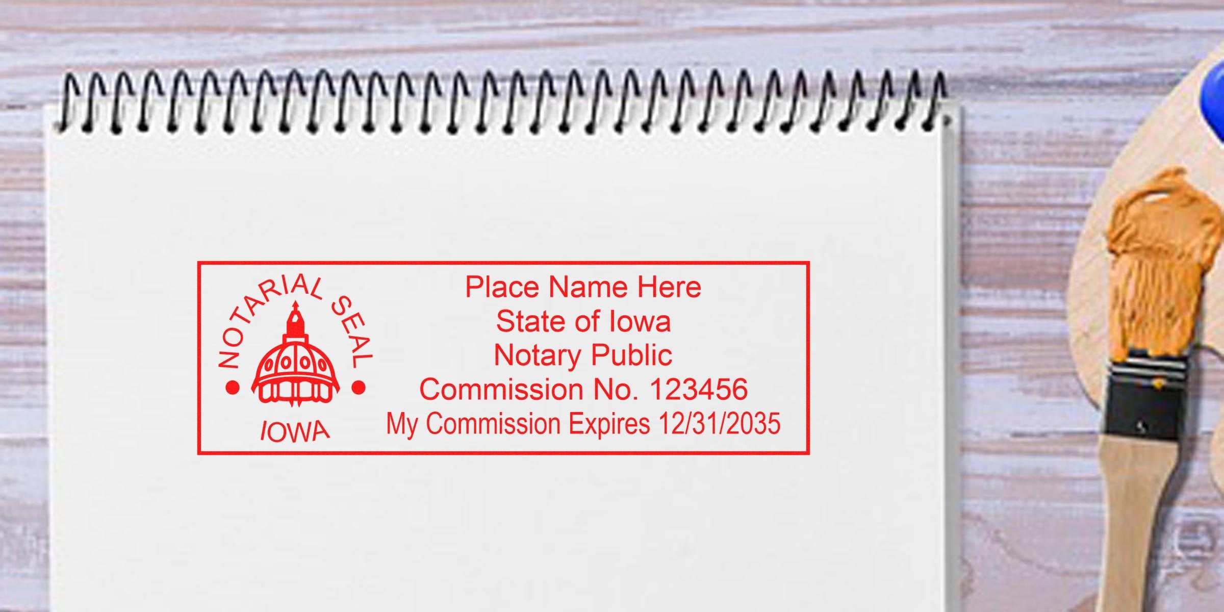 Red Iowa notarial seal rubber stamp impression on white spiral notebook showing sample notary public text and commission details, on a light wooden table with a paintbrush nearby.