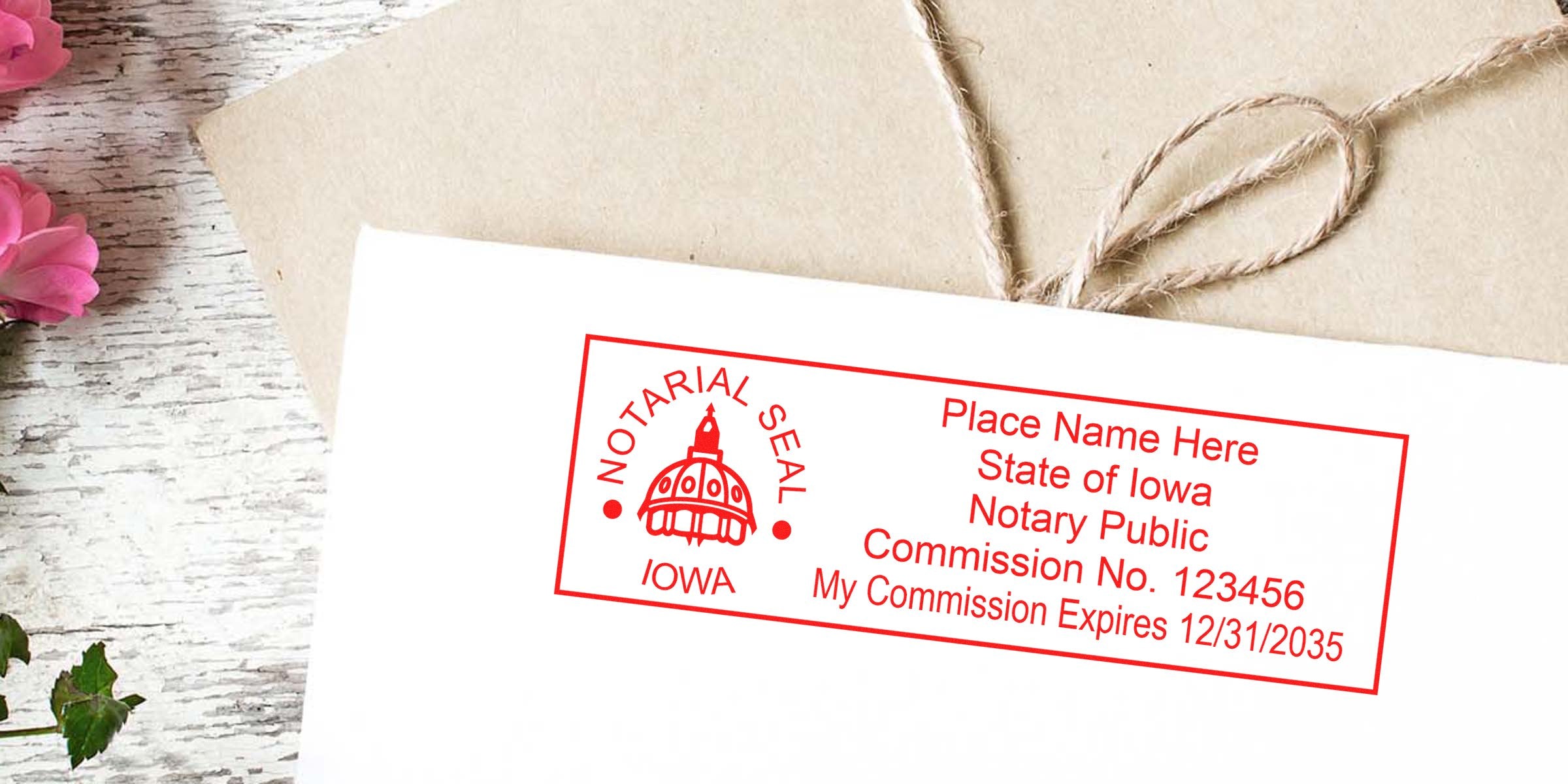 Red Iowa notary public stamp on white envelope showing capitol dome logo, sample commission number and expiration date; kraft paper envelope with twine and pink flowers on rustic white wood background.