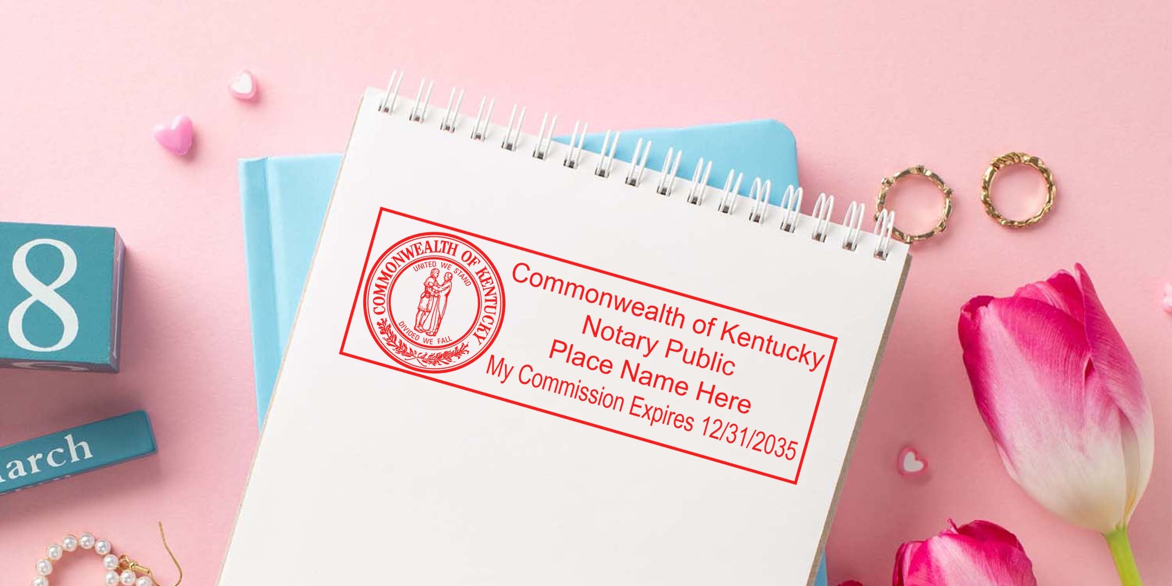 Overhead flatlay of spiral notebook stamped with red Commonwealth of Kentucky notary public seal on pink desk with tulips, gold rings, pearl bracelet and blue calendar blocks