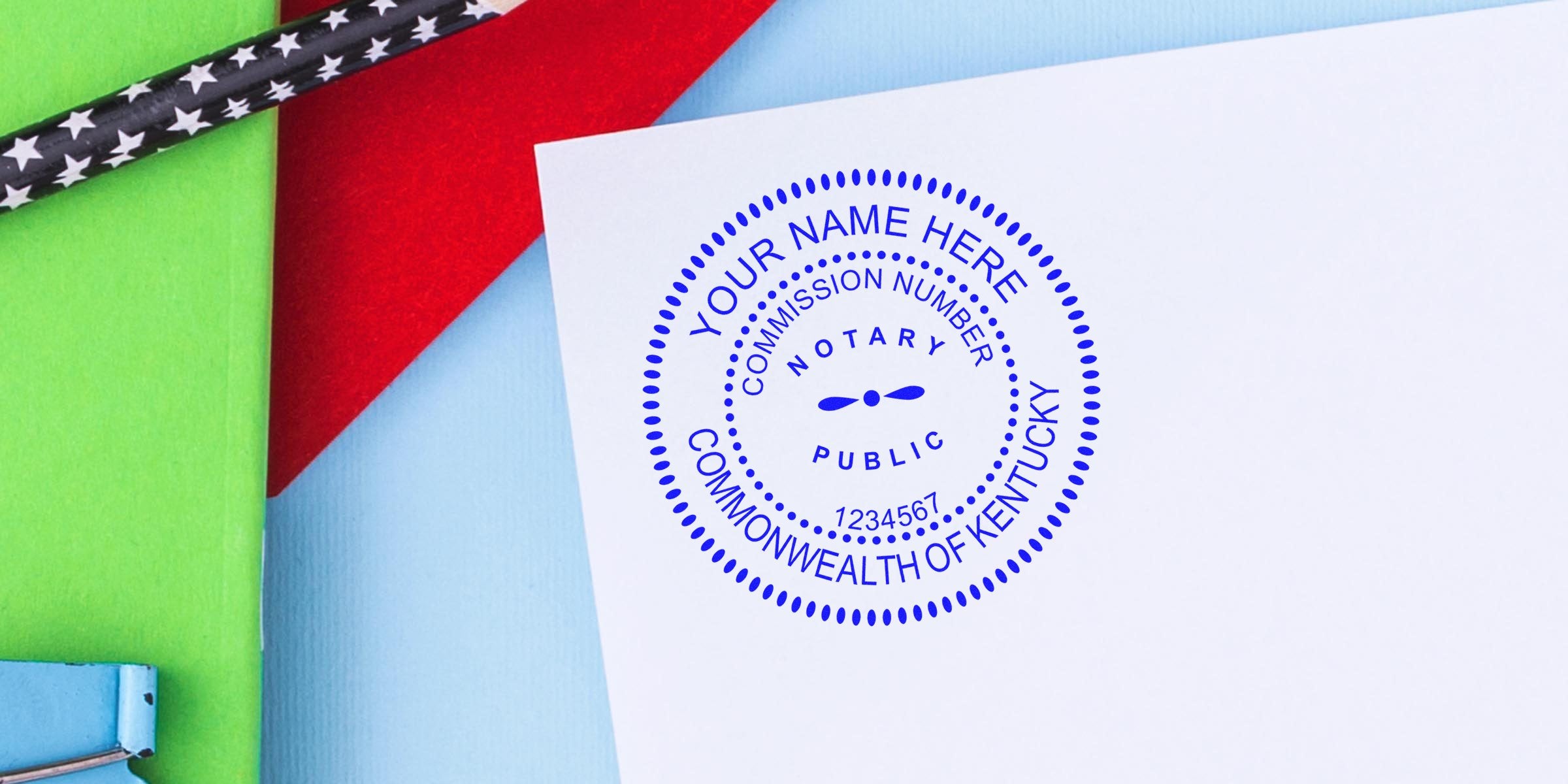 Blue circular notary stamp reading "Notary Public" and "Commonwealth of Kentucky" with commission number on white paper, photographed on colorful background with green and red folders and star-pattern pencil