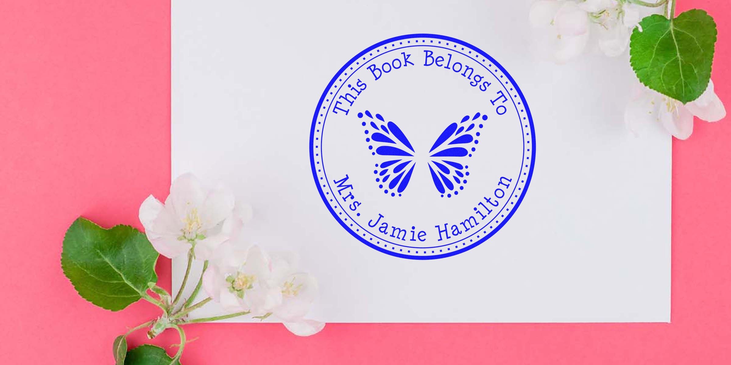 Blue circular stamp on a white sheet that reads “This Book Belongs To Mrs Jamie Hamilton” with a butterfly graphic, set on a pink background with white flowers.