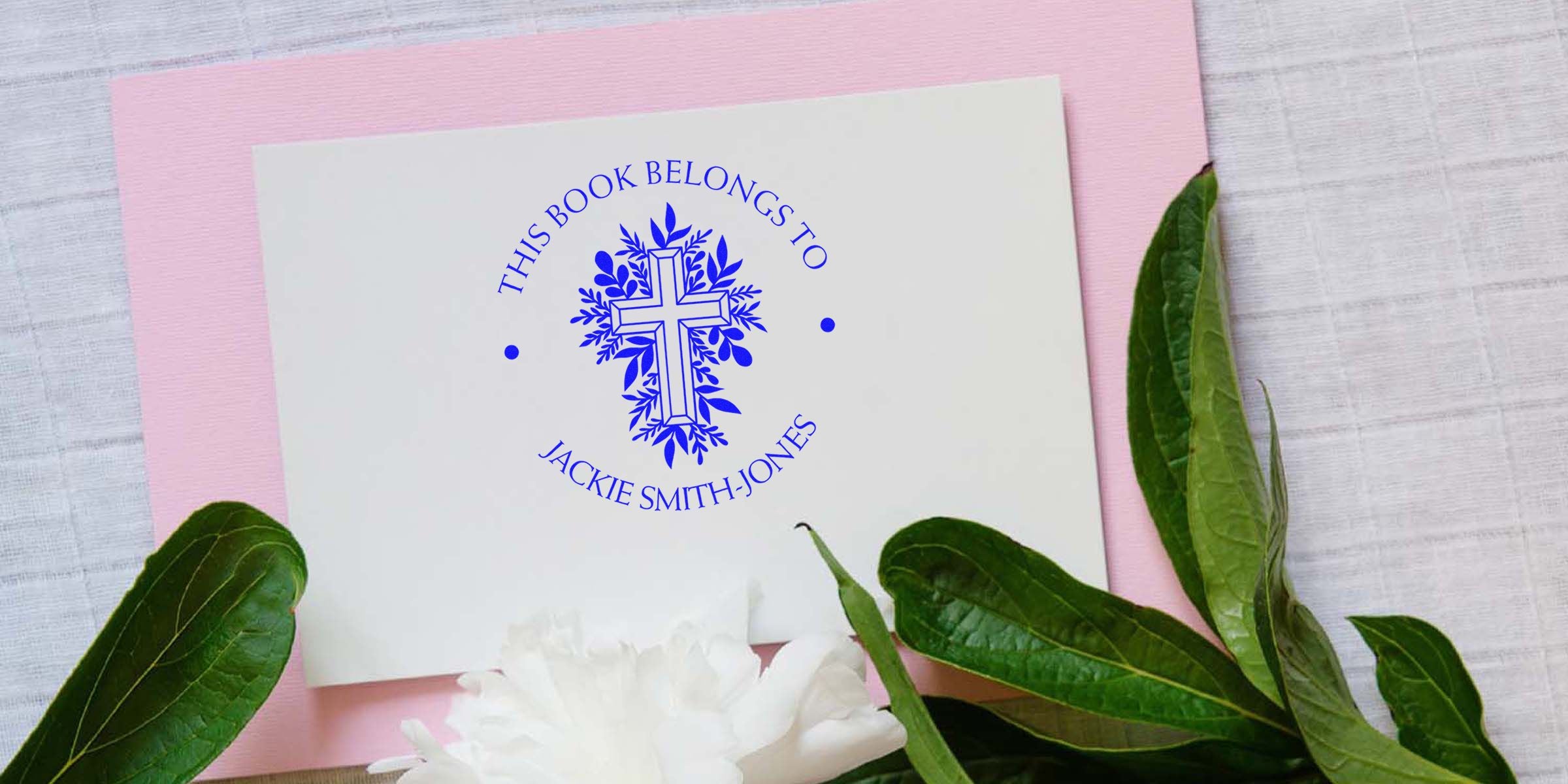 Card with blue decorative cross and the text “THIS BOOK BELONGS TO JACKIE SMITH-JONES” on a white surface, set on a pink background with green leaves nearby.