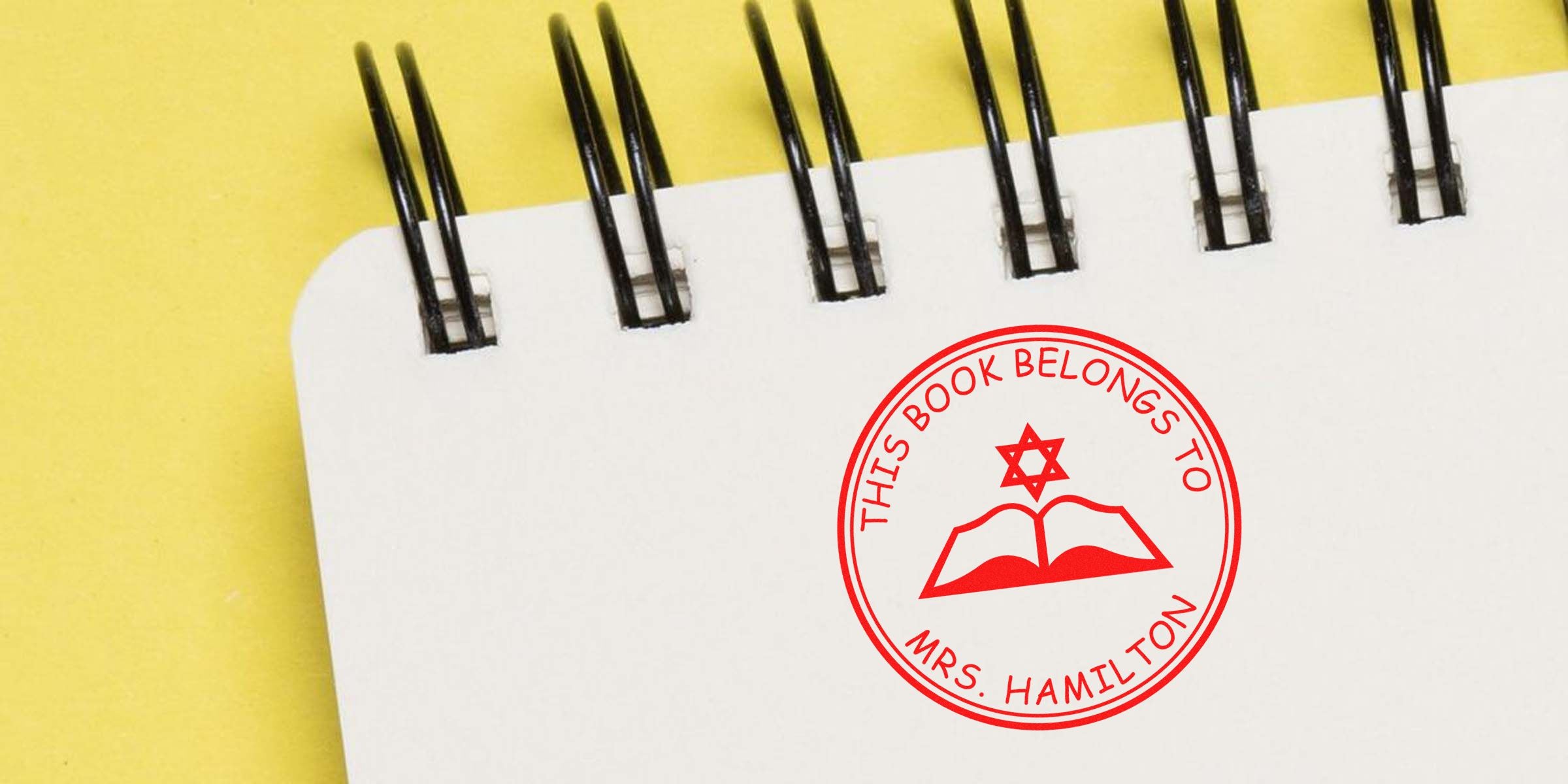 Red circular stamp reading “This Book Belongs To Mrs. Hamilton” with a star of David above an open book icon on a white notebook page.