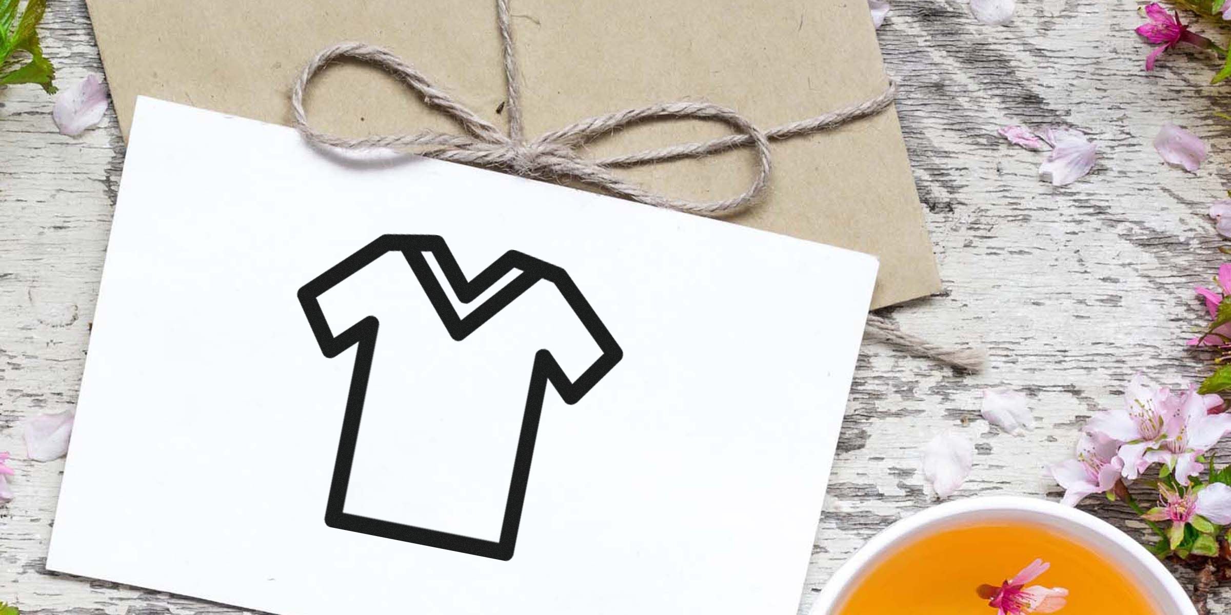 White card with a black outline of a T-shirt on a rustic wooden table, with twine, pink flowers, and an orange cup nearby.