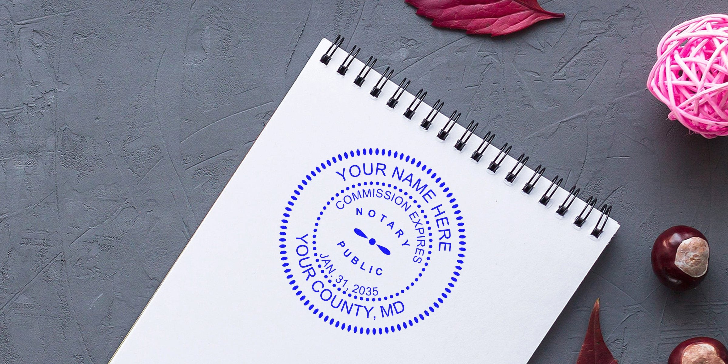Top-down view of a spiral notebook page with a blue notary stamp mockup reading "Your Name Here, Your County, MD" on a textured gray background with decorative pink rattan ball and chestnuts