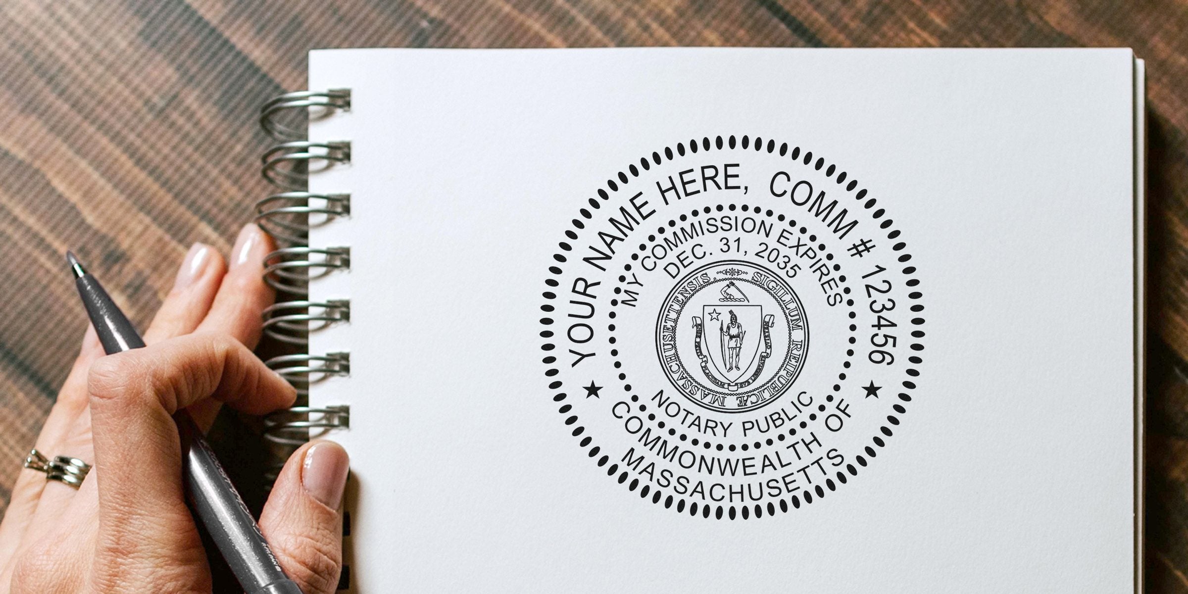 Close-up of a hand holding a pen over a spiral notebook featuring a black Massachusetts notary public seal stamp design on white paper.