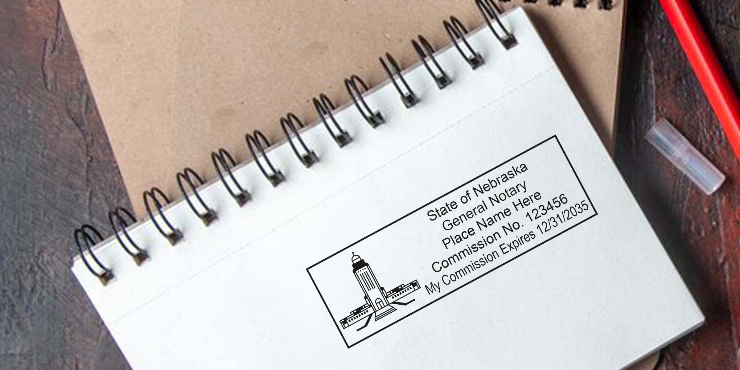 Spiral notebook page with a printed Nebraska notary stamp showing state capitol icon, sample notary text, commission number and expiry date; brown cover beneath and red pen cap at right, flat lay office stationery image.