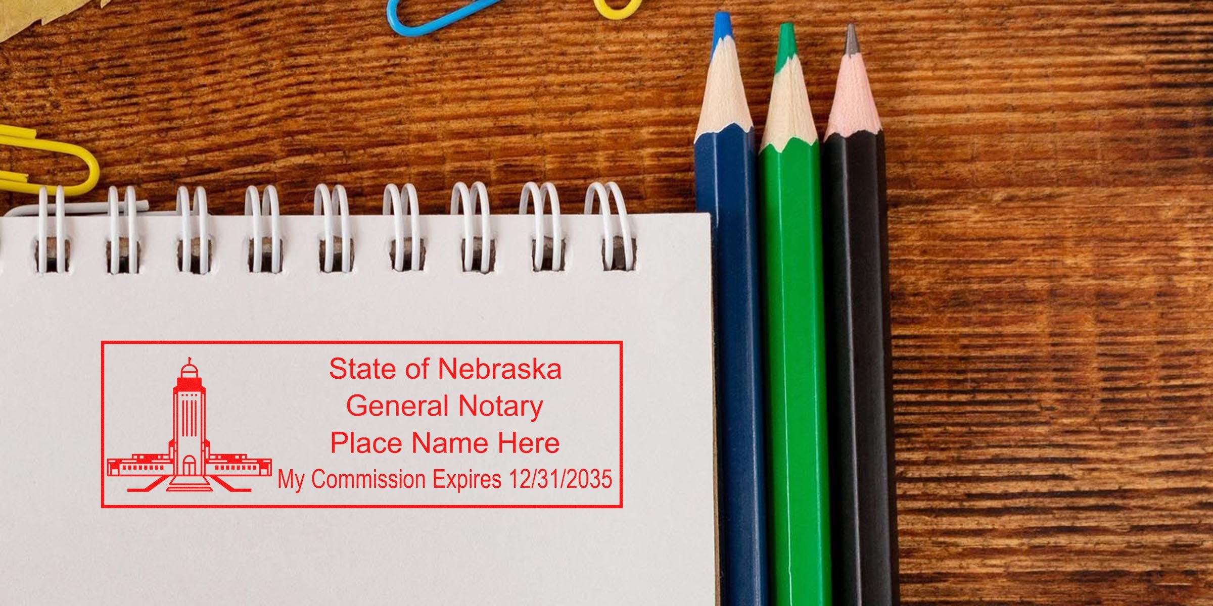 Spiral notebook with red "State of Nebraska General Notary" stamp mockup showing placeholder name and commission expiry, three colored pencils (blue, green, black) and paperclips on a wooden desk — notary stamp stationery concept