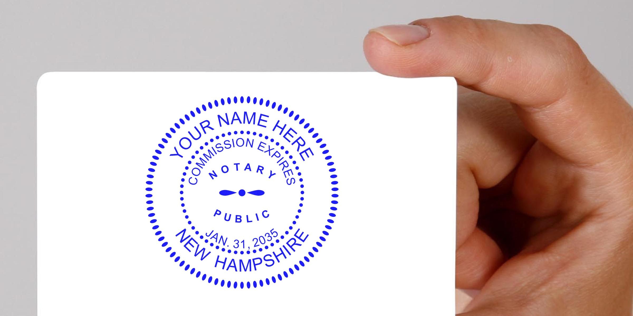 Close-up of a hand holding a white card displaying a blue circular New Hampshire notary public seal with dotted border, placeholder "Your Name Here," commission expires Jan. 31, 2035.