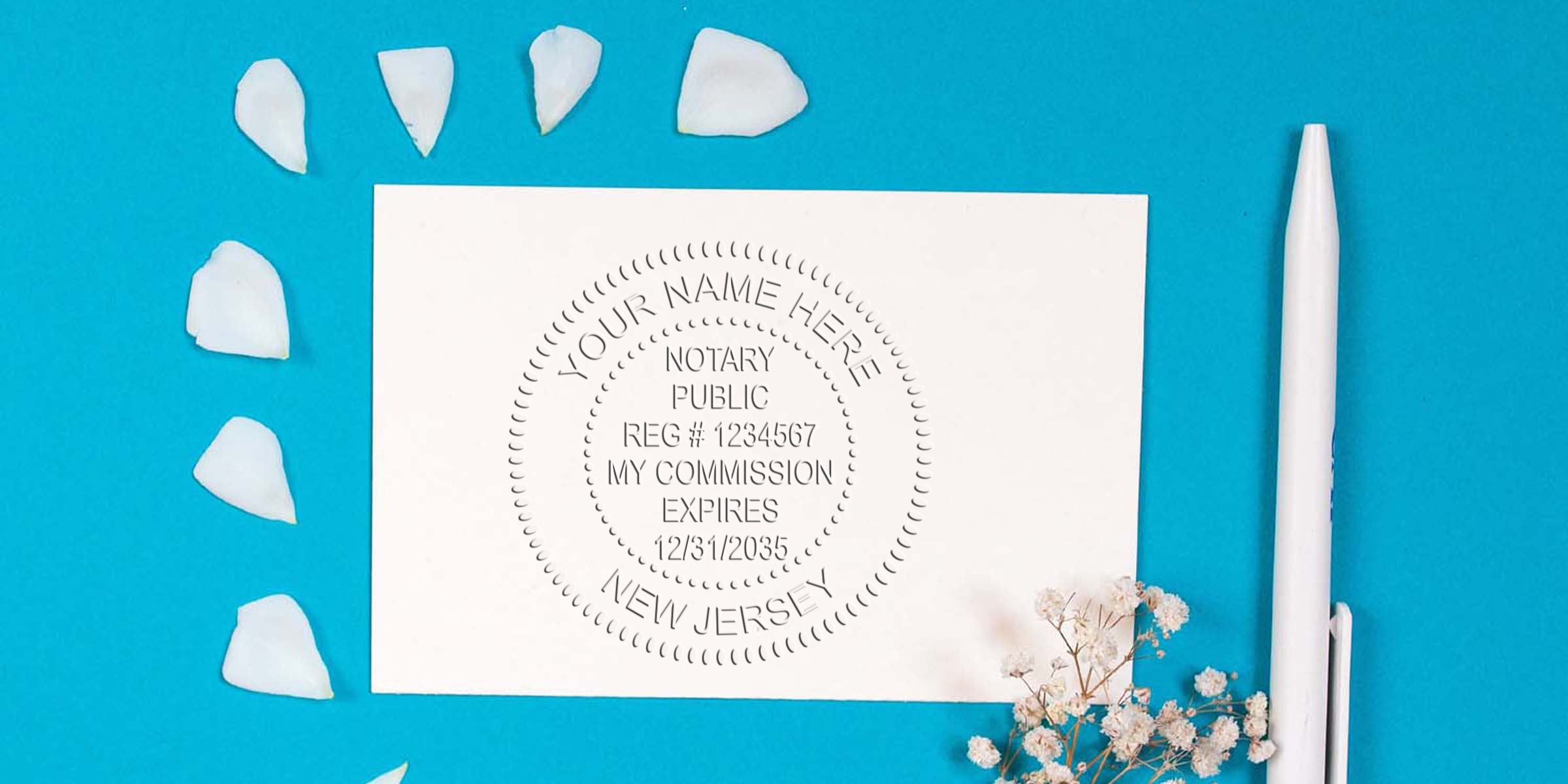 Flat-lay of a customizable New Jersey notary seal sample on white card with registration number and commission expiry text, surrounded by white petals, small flowers and a white pen on blue background