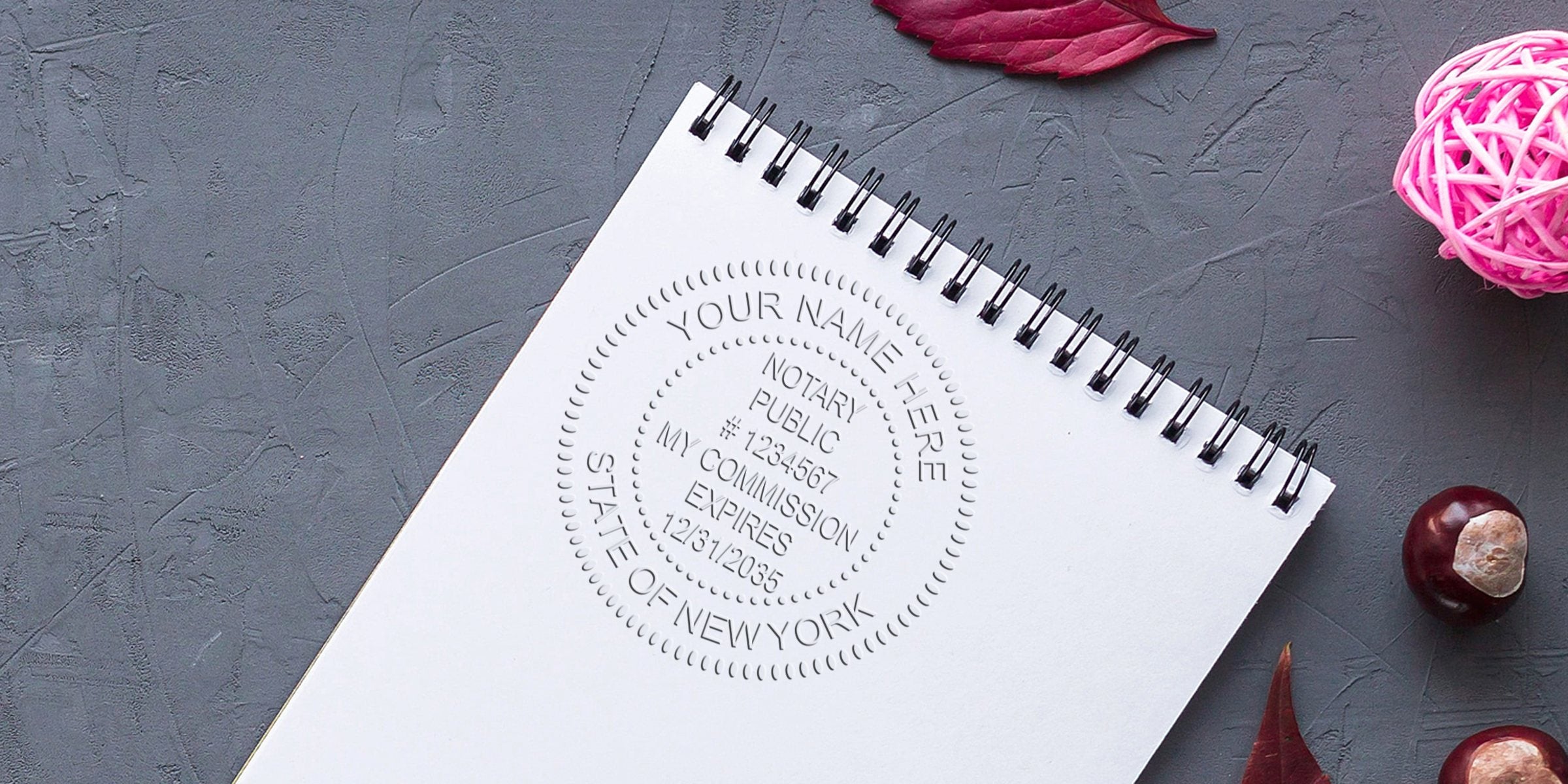 Flat-lay spiral notebook with embossed New York notary public seal on gray textured background, decorative pink rattan ball and autumn leaves, close-up of notary stamp impression for legal documents and certification.