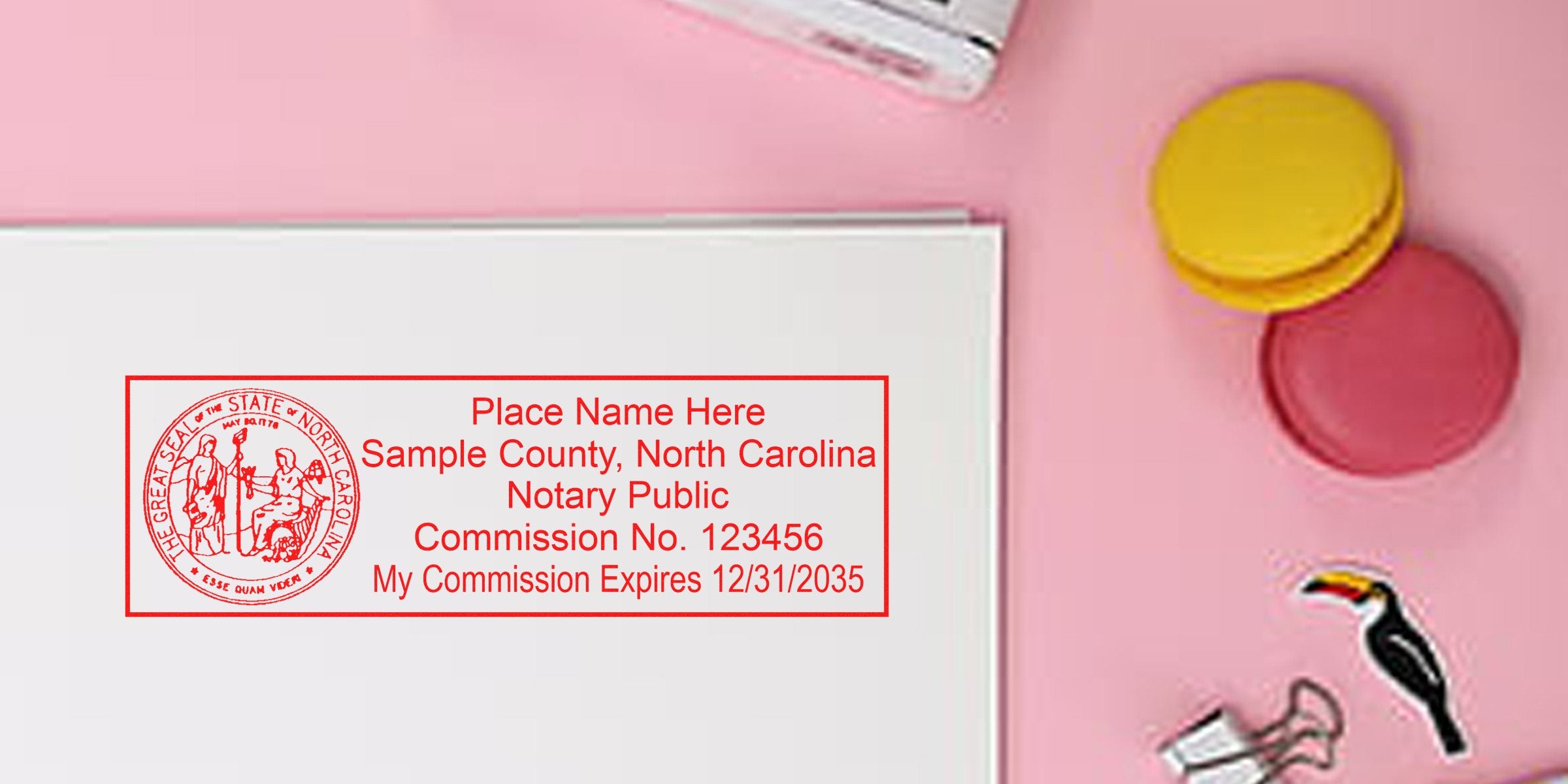 Red rectangular North Carolina notary public stamp sample in red ink on white paper, placed on pink desk with yellow and pink macarons and binder clip