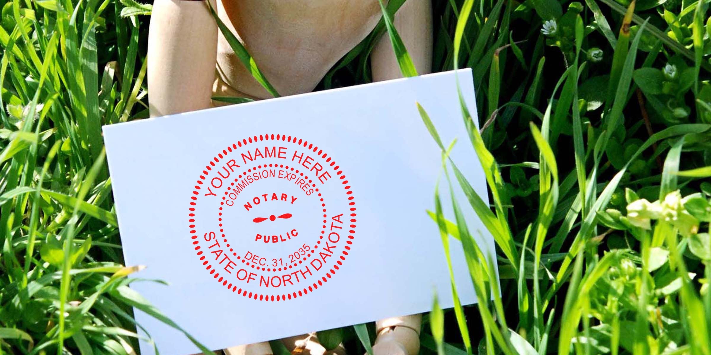 White envelope with red circular "Notary Public, State of North Dakota" seal held among green grass blades, outdoor close-up