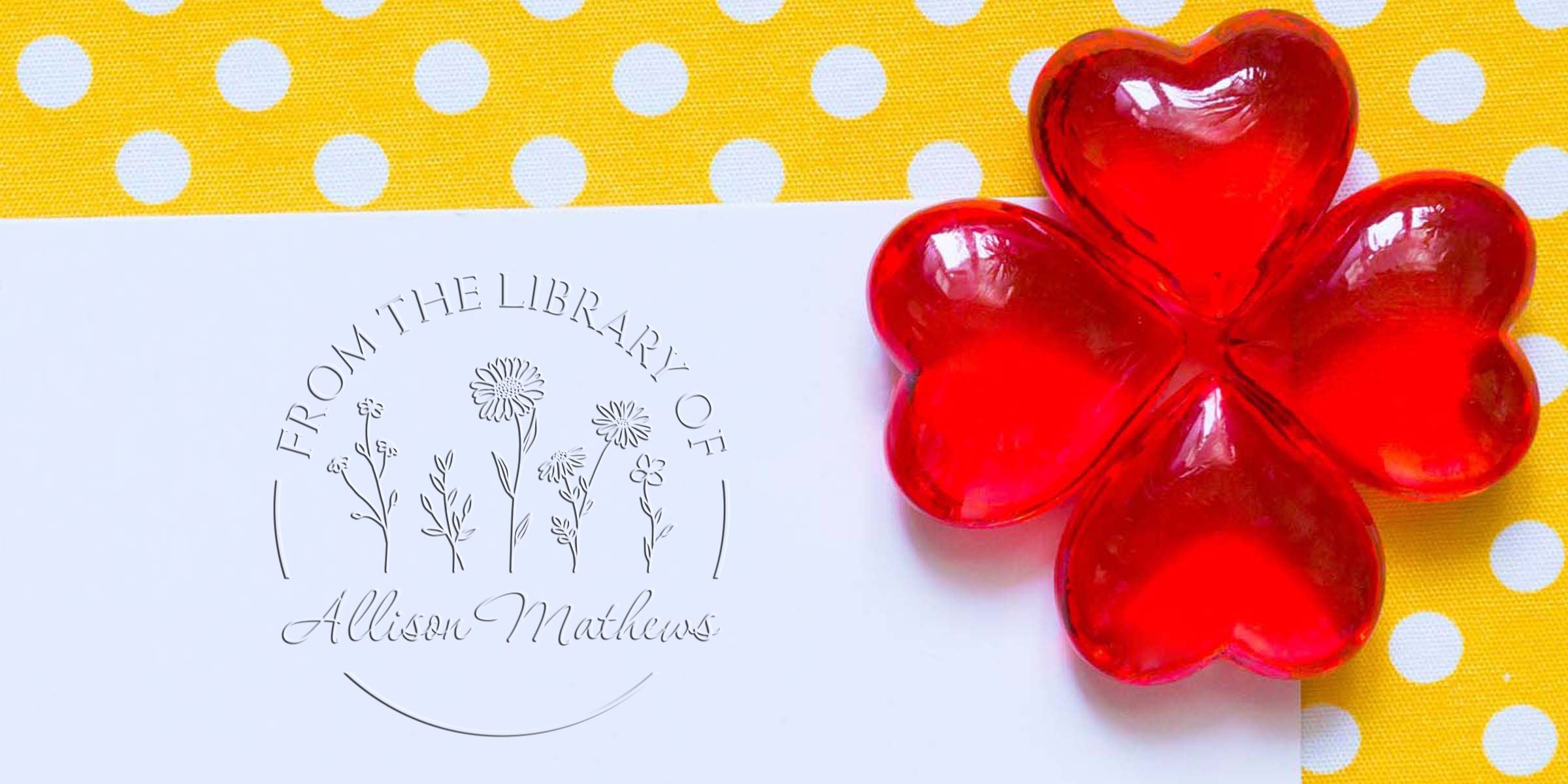 A white card with a circular logo reading “From the Library of Allison Mathews” beside a bright red glass flower on a yellow polka-dotted background.