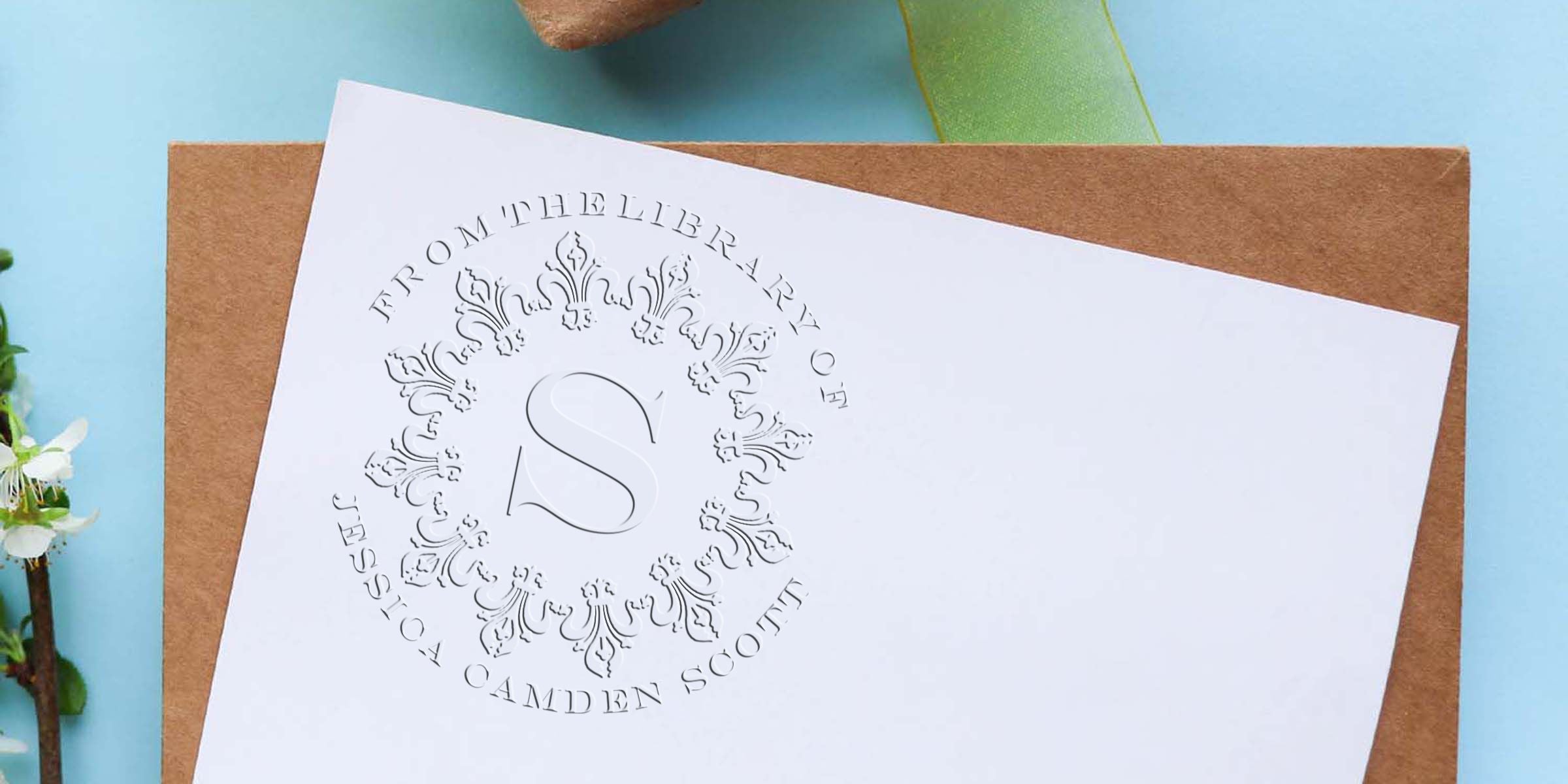 Embossed white card with an ornate circular frame and a large “S” in the center, placed on a brown envelope.