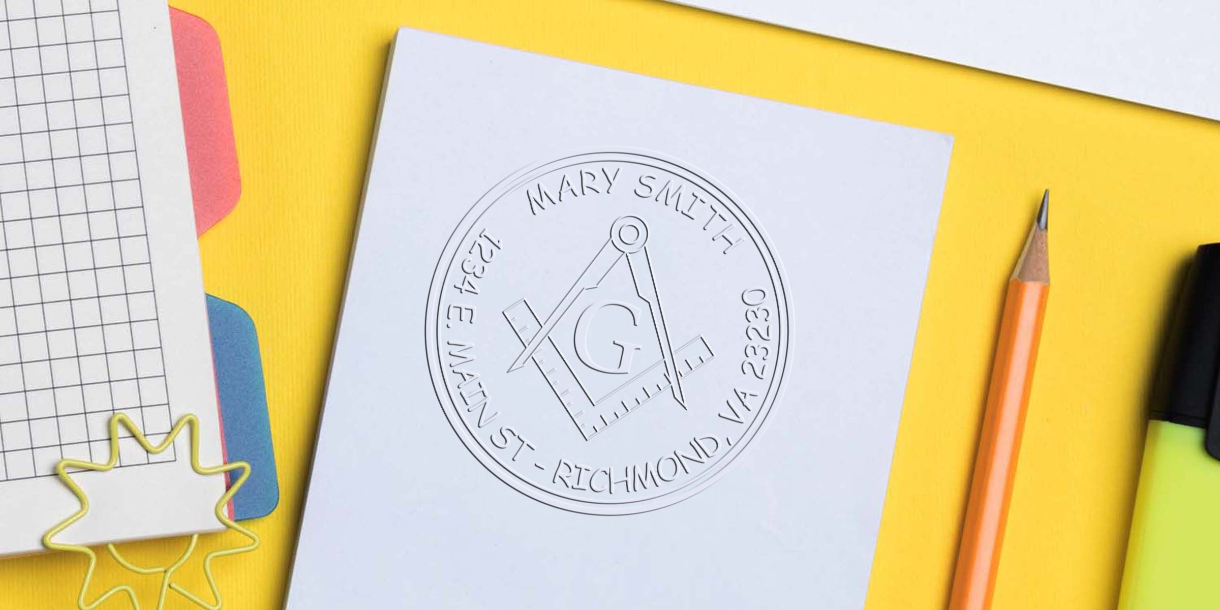 A white square card featuring an embossed Masonic square and compasses symbol with a central G, surrounded by circular text, on a bright yellow background with a pencil and highlighter nearby.