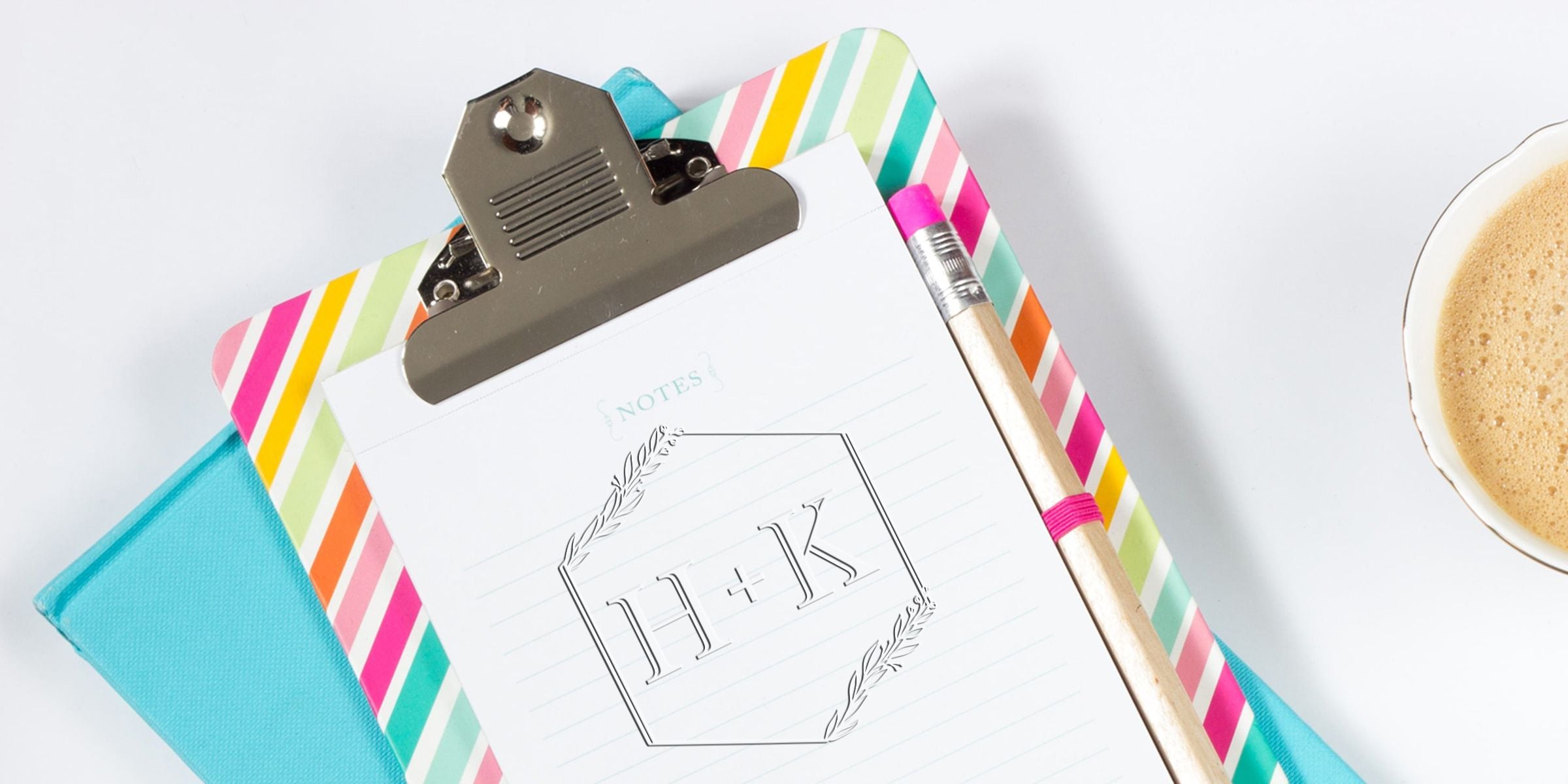 Clipboard with a sheet showing an H+K logo inside a hexagon, surrounded by colorful striped borders, a pencil, and a cup of coffee on the right.