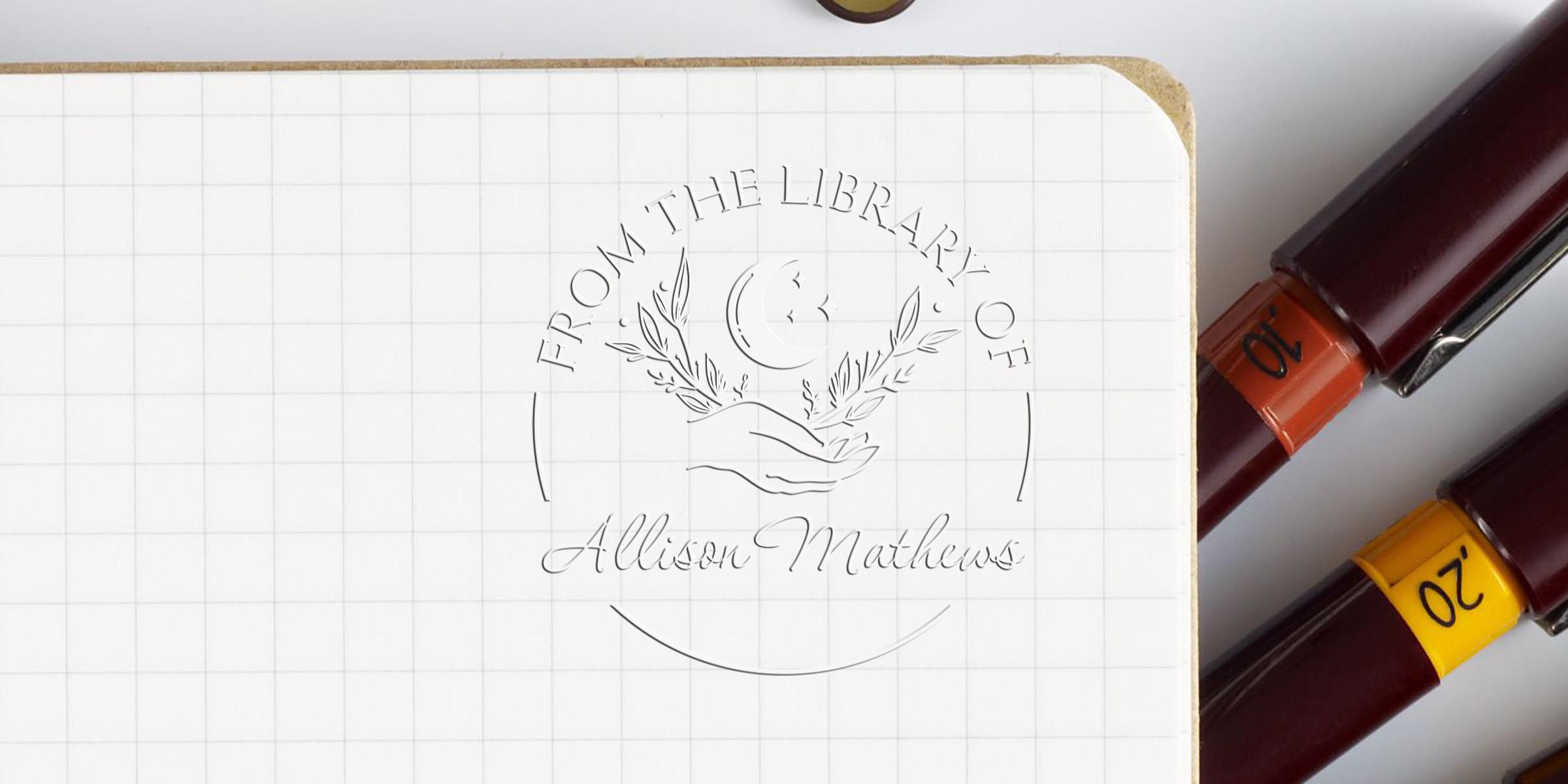 Notebook page featuring an embossed circular logo reading “From the Library of Allison Mathews” with two ink pens (orange and yellow caps) resting alongside.
