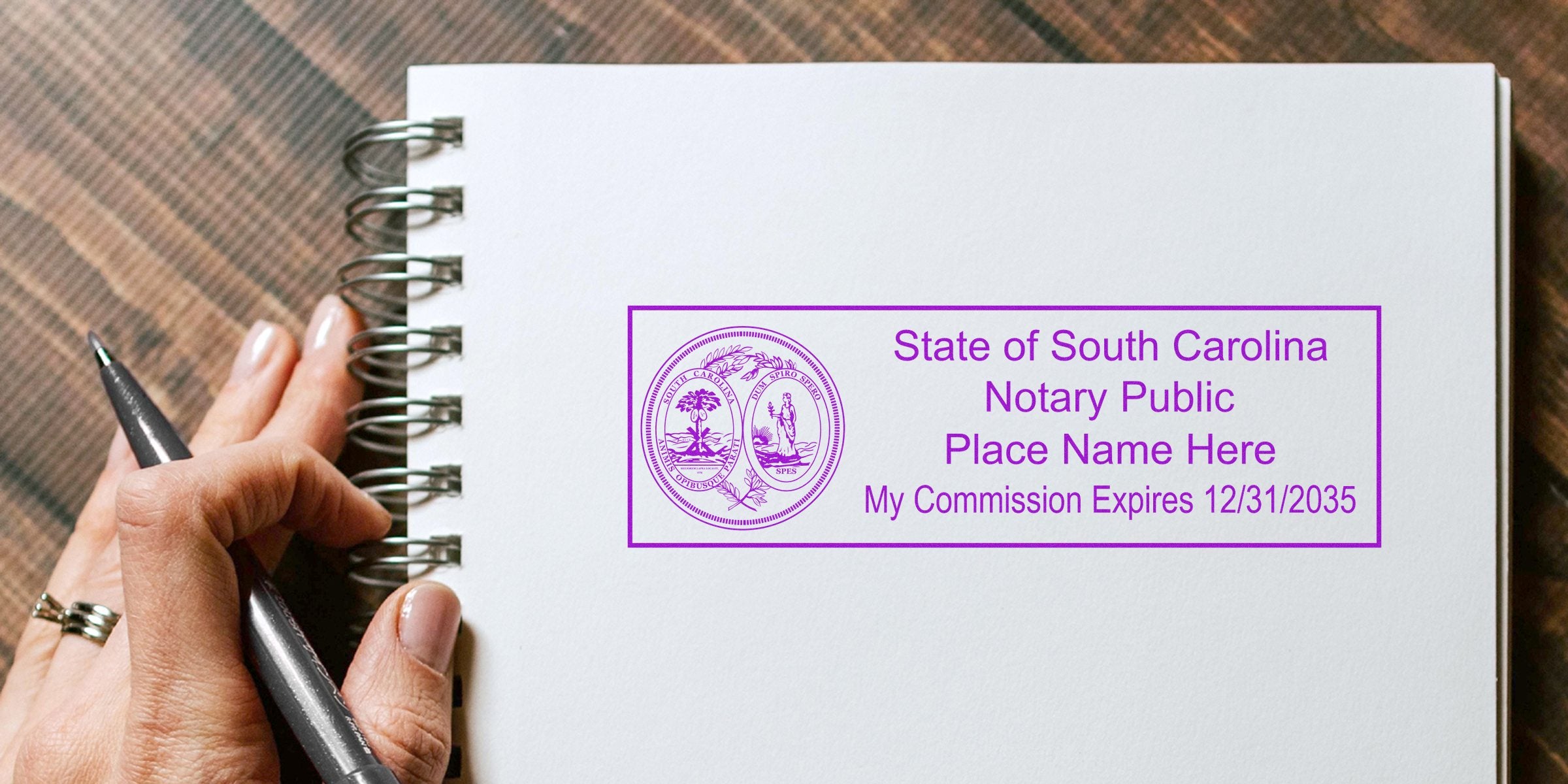 Purple State of South Carolina notary public stamp reading "Place Name Here" and "My Commission Expires 12/31/2035" on blank spiral notebook page with a hand holding a pen nearby.