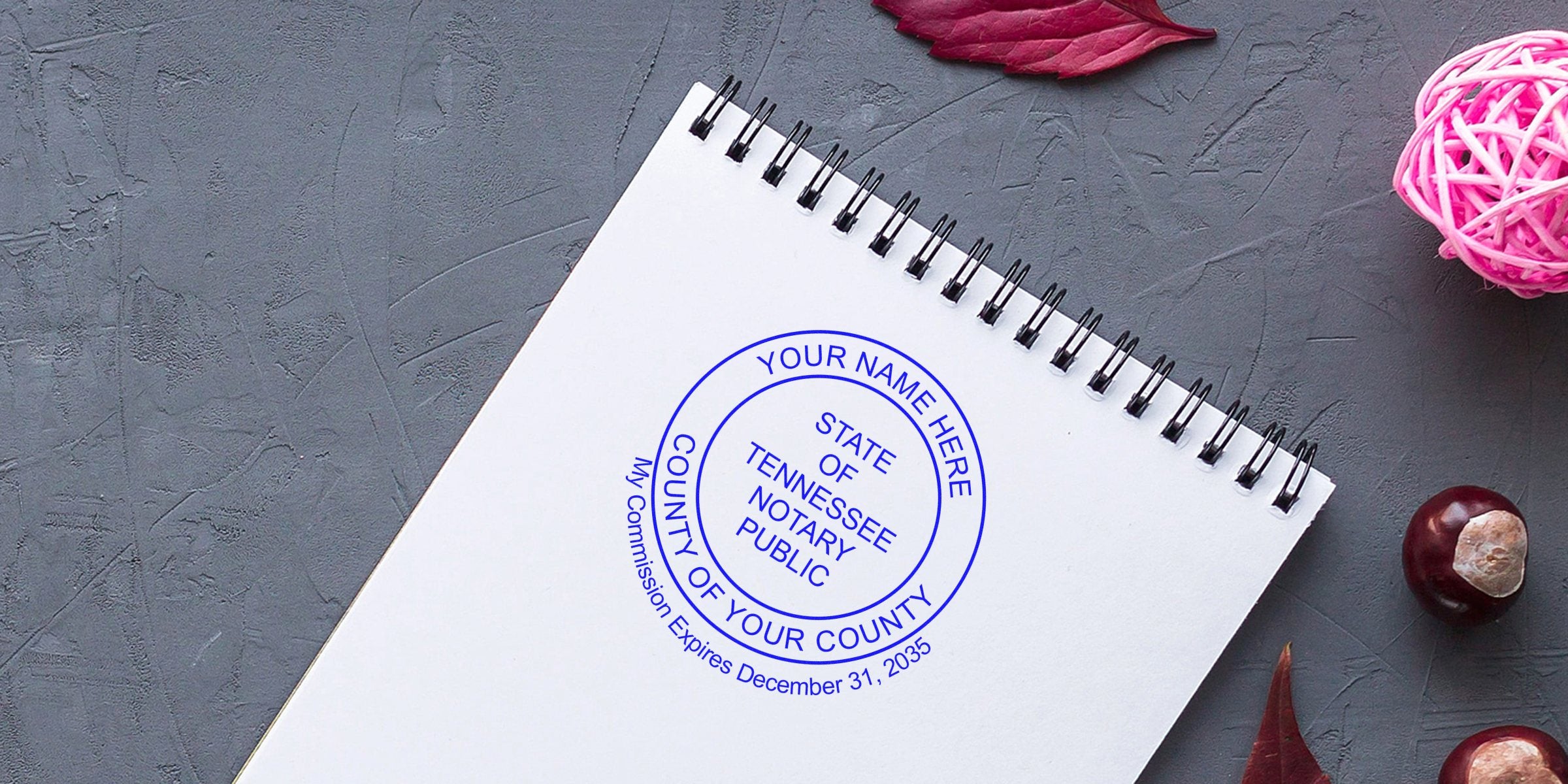 Spiral notebook with blue circular "State of Tennessee Notary Public" stamp mockup on textured gray background, pink decorative ball and chestnuts, flat lay composition