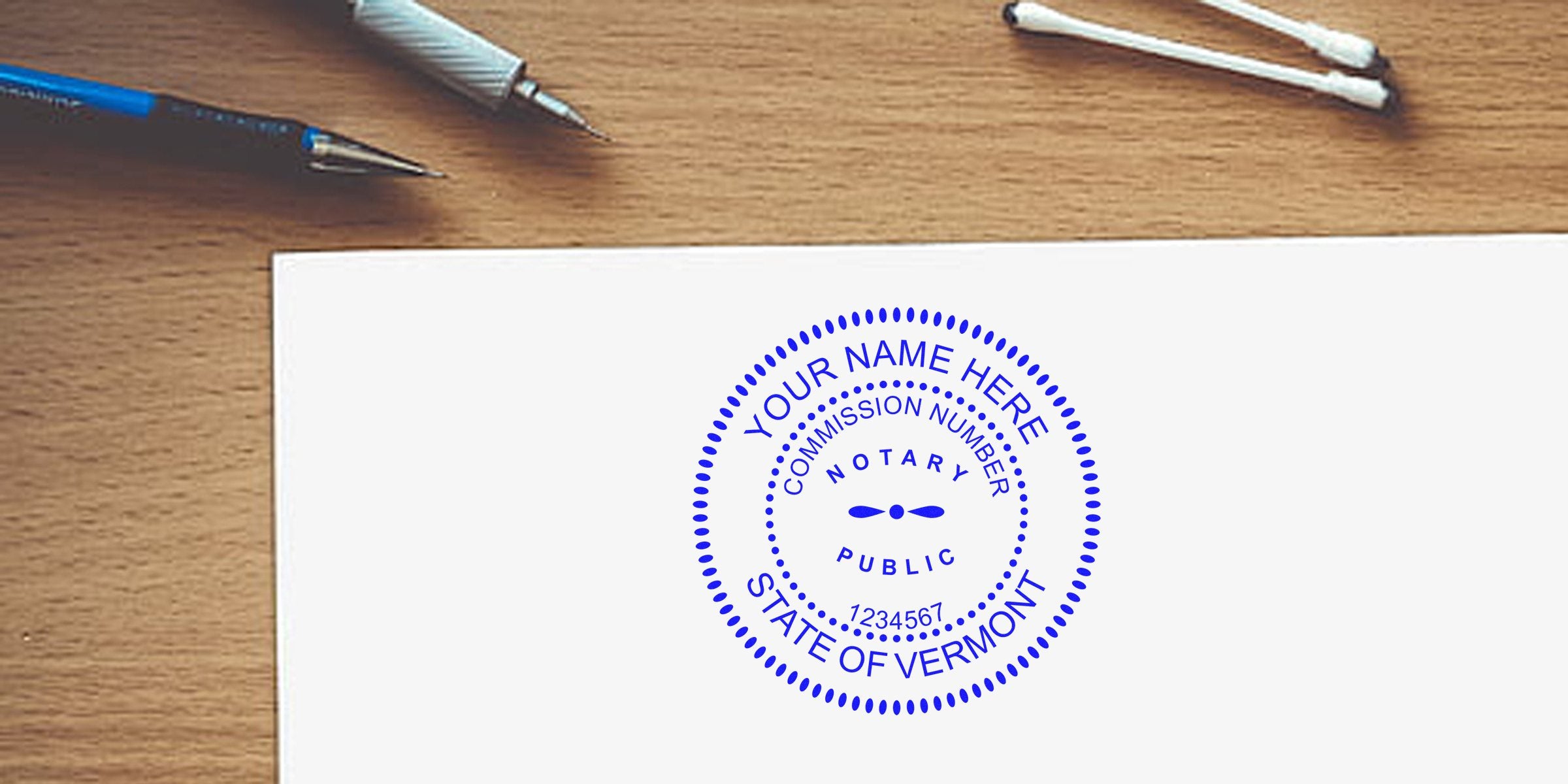 Blue circular "Notary Public" stamp mockup reading "State of Vermont" and commission number on white paper at a wooden desk with pens and cotton swabs.