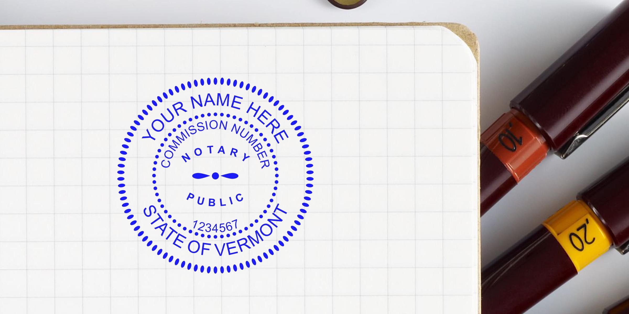 Blue circular notary public stamp mockup reading "Your Name Here," "State of Vermont" and sample commission number on graph-paper notebook beside two fountain pens.