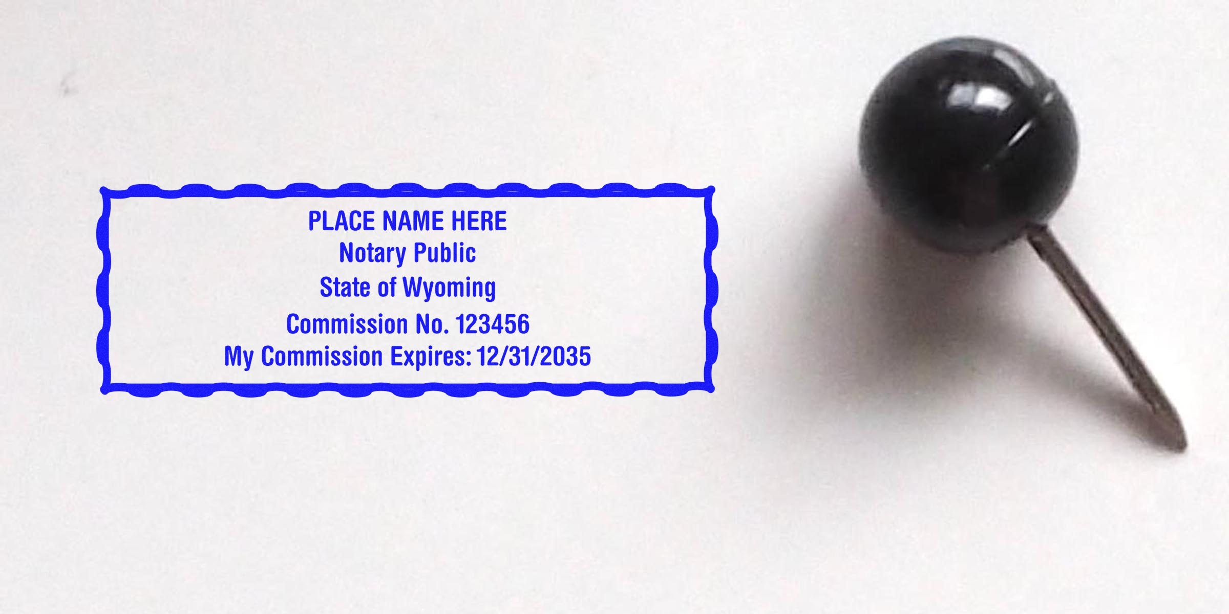 Blue rectangular notary stamp sample reading "PLACE NAME HERE Notary Public State of Wyoming Commission No. 123456 My Commission Expires: 12/31/2035" beside a black push pin on white paper