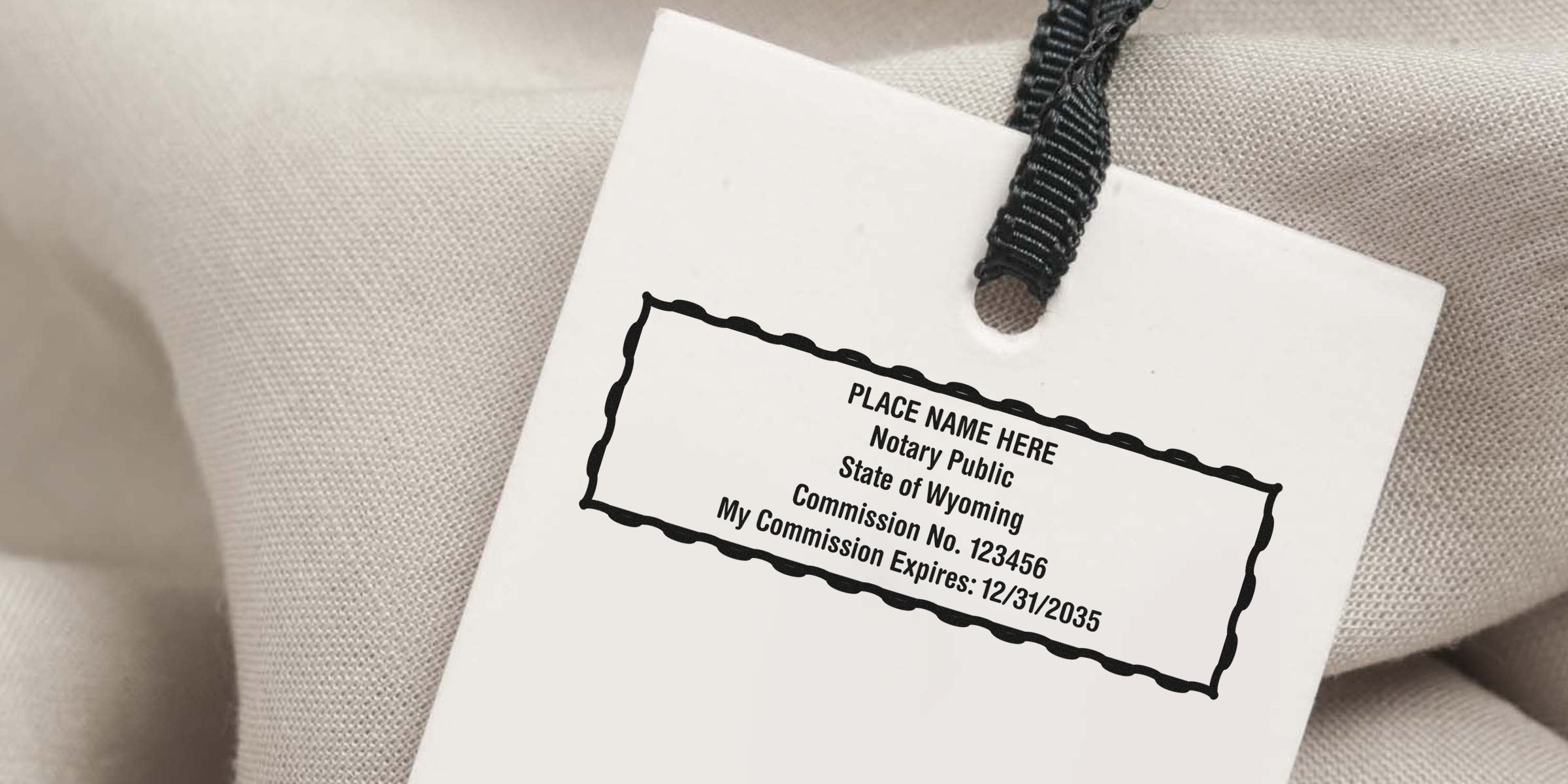 White clothing hang tag with black notary stamp reading "Place Name Here Notary Public State of Wyoming Commission No. 123456 My Commission Expires: 12/31/2035," attached by a black ribbon on beige textured fabric background.