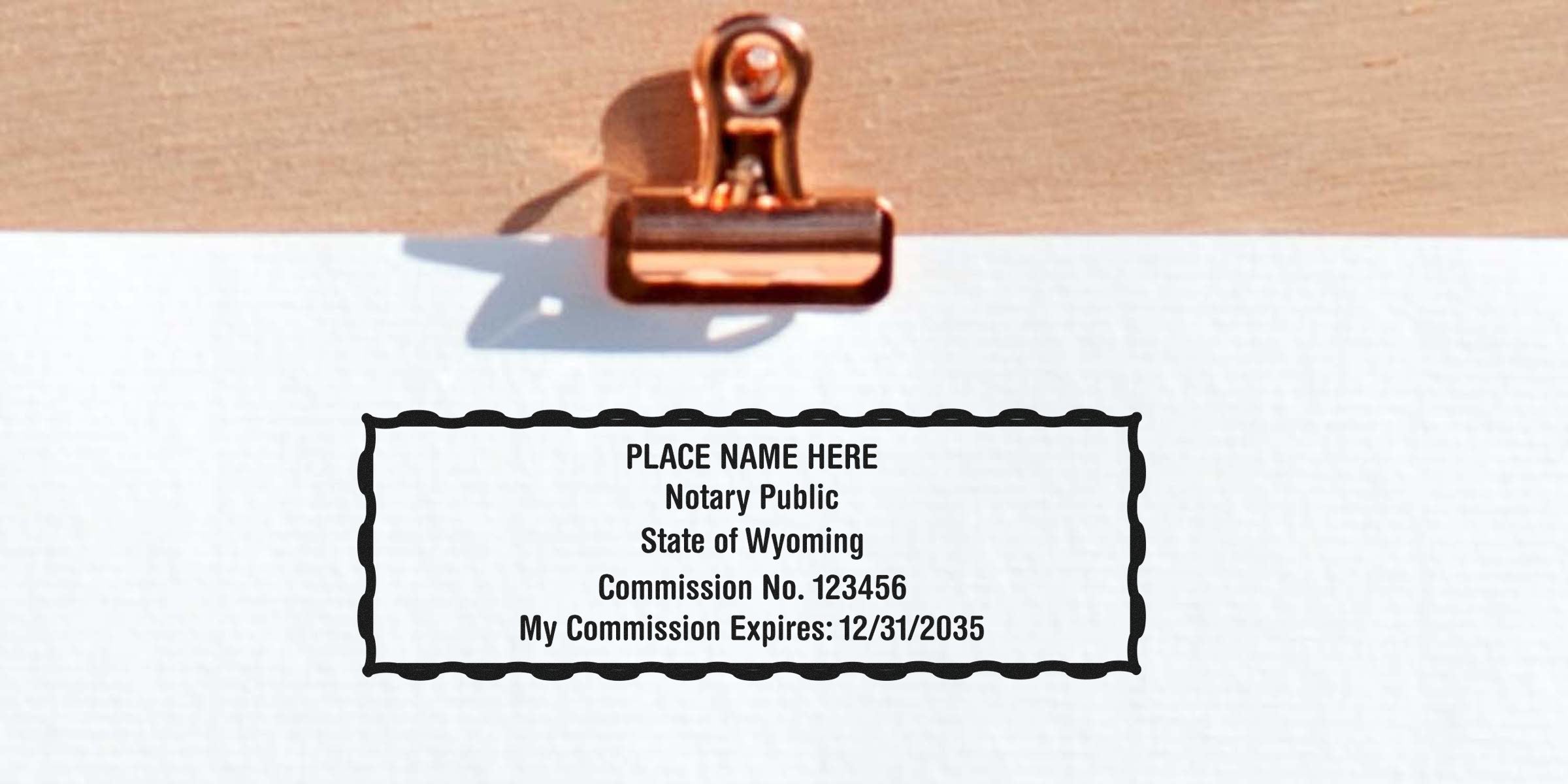 Rose gold binder clip holding white paper with a black notary stamp template reading "PLACE NAME HERE, Notary Public, State of Wyoming, Commission No. 123456, My Commission Expires: 12/31/2035".