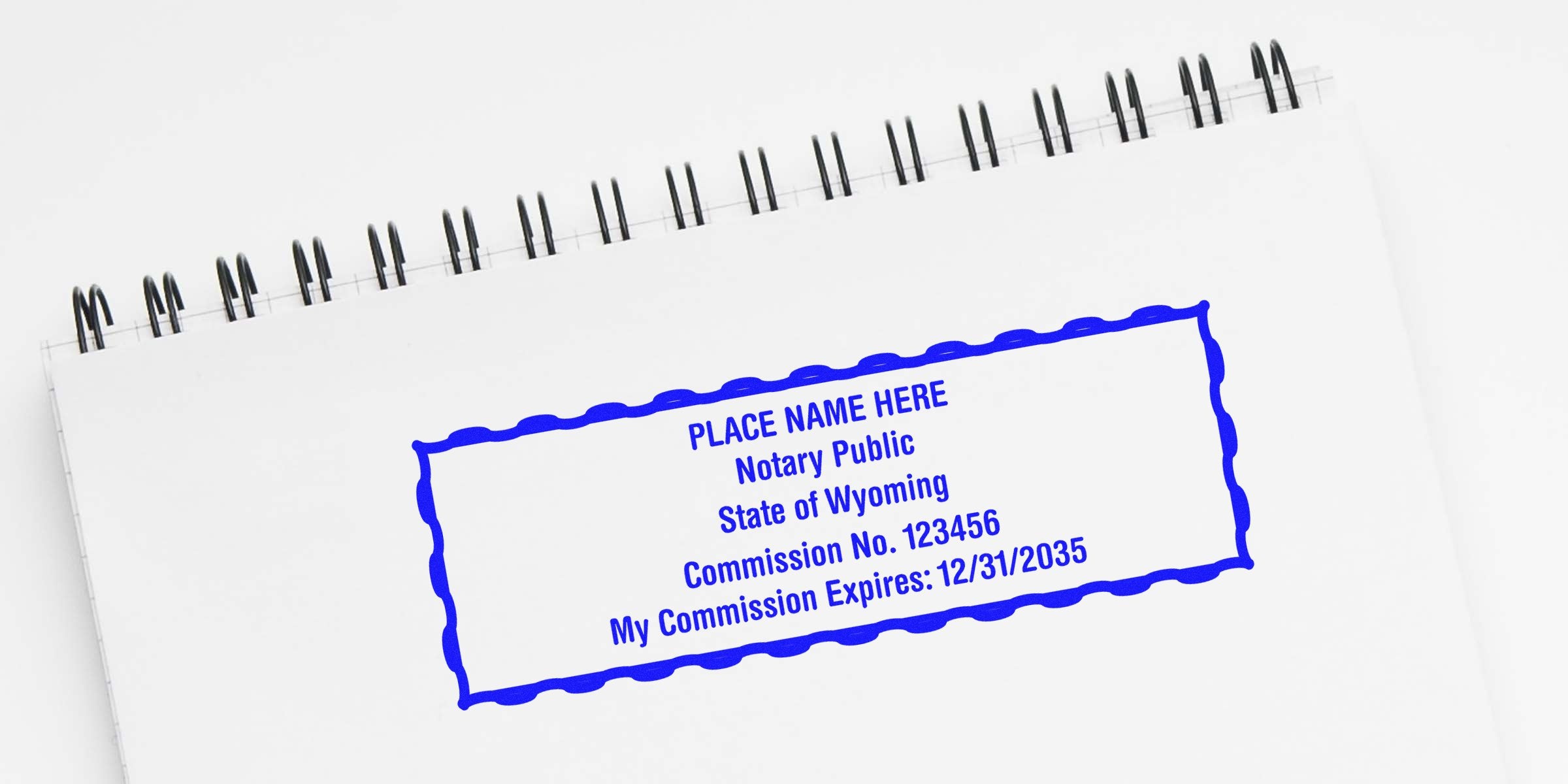 Blue rectangular notary stamp on a spiral-bound white notebook reading "PLACE NAME HERE Notary Public State of Wyoming Commission No. 123456 My Commission Expires: 12/31/2035" with a wavy decorative border.