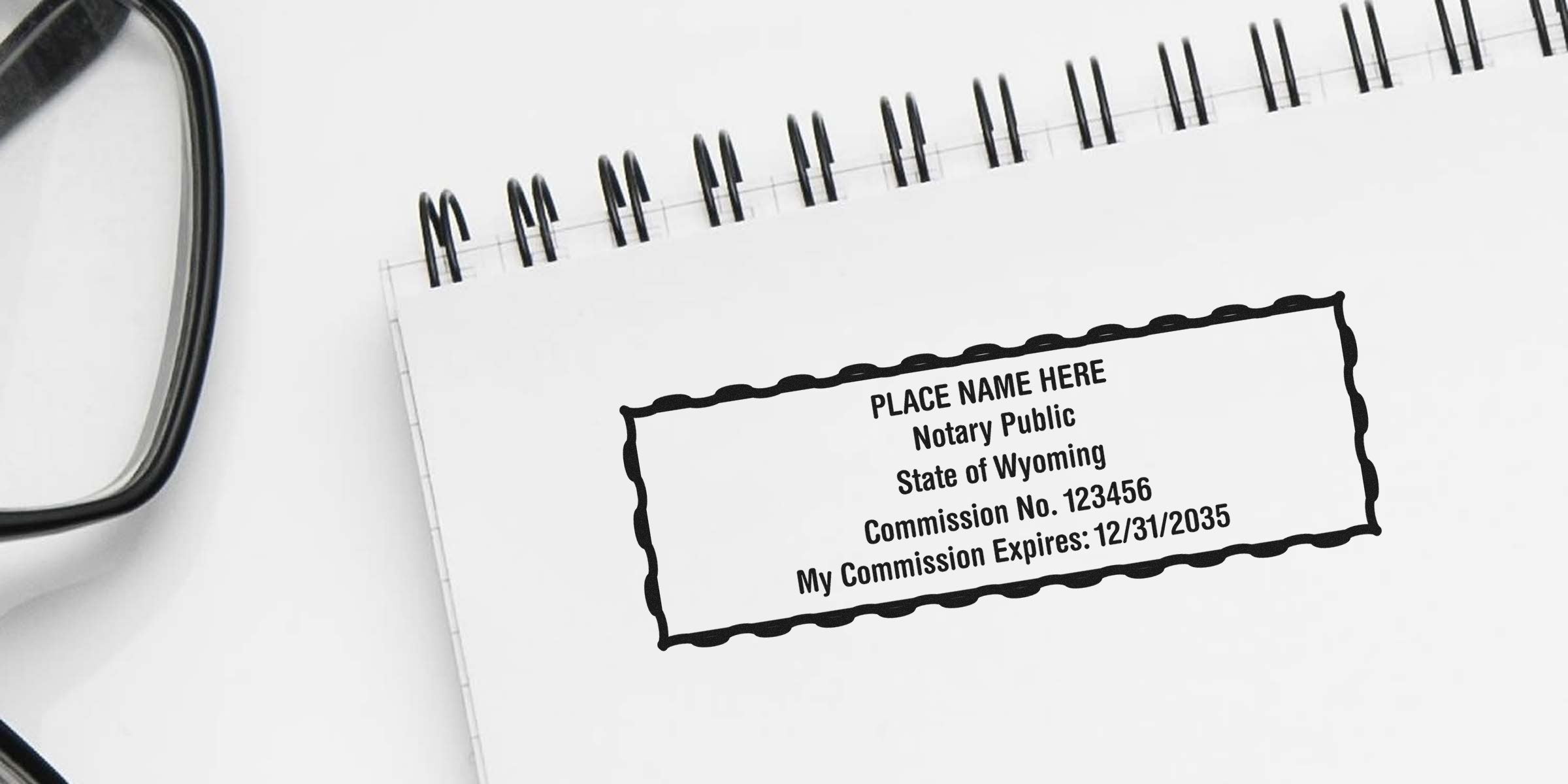 Spiral notebook page with a sample Wyoming notary stamp reading "PLACE NAME HERE Notary Public State of Wyoming Commission No. 123456 Expires 12/31/2035" and partial black eyeglasses frame.