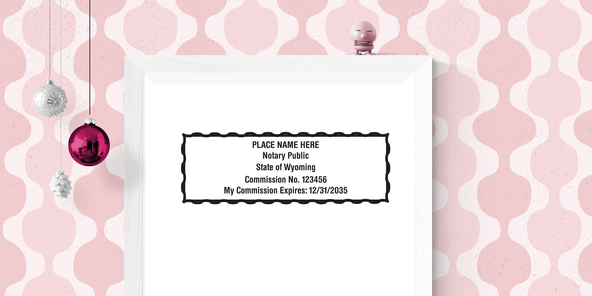 Mockup notary stamp template in white frame reading "PLACE NAME HERE Notary Public State of Wyoming Commission No. 123456" on pink retro wallpaper with hanging ornaments