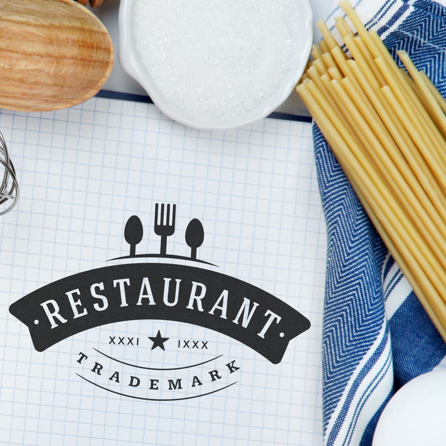 Business Stamps with a Logo Blog Post Feature Image: A custom restaurant stamp on graph paper, surrounded by pasta, salt, and a kitchen towel.